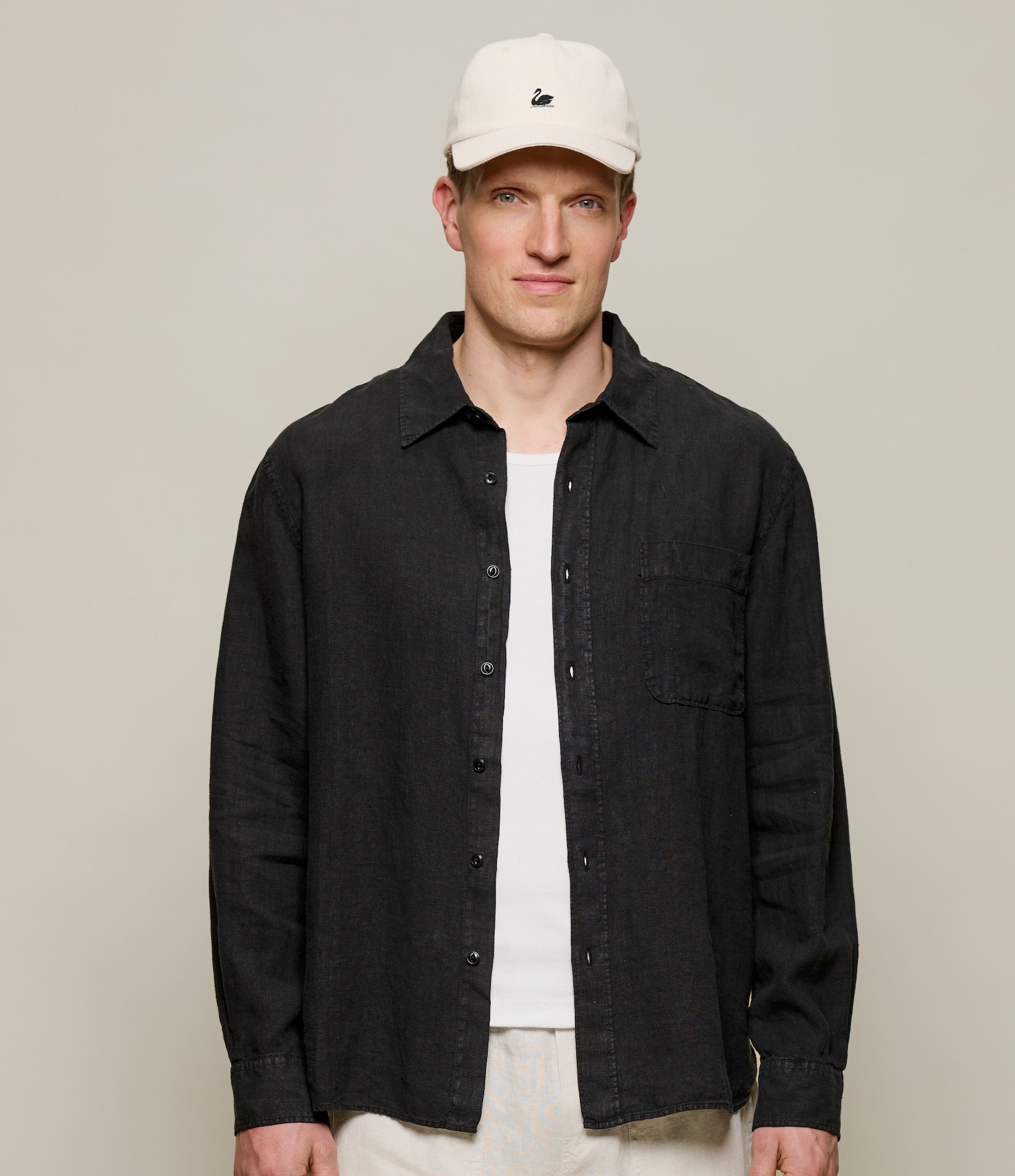 Man wearing a black linen shirt, white t-shirt, and beige cap against a plain background