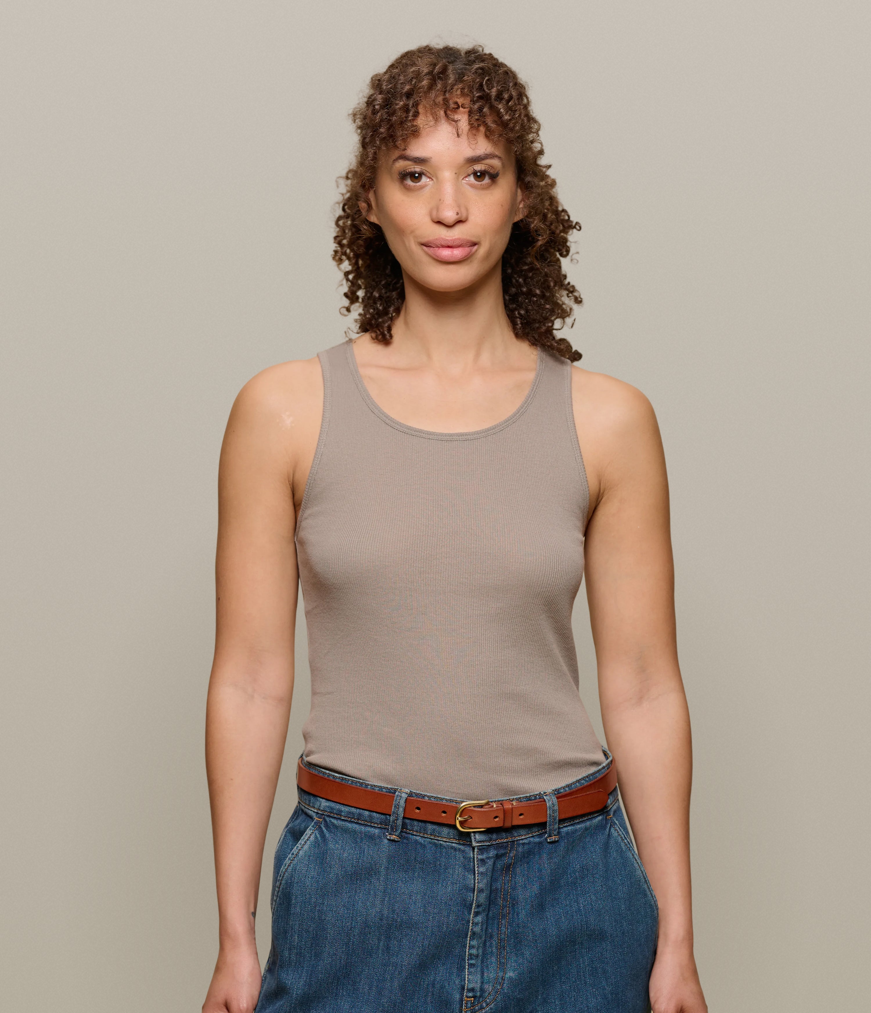 Woman wearing a brown ribbed tank top and blue jeans against a beige background