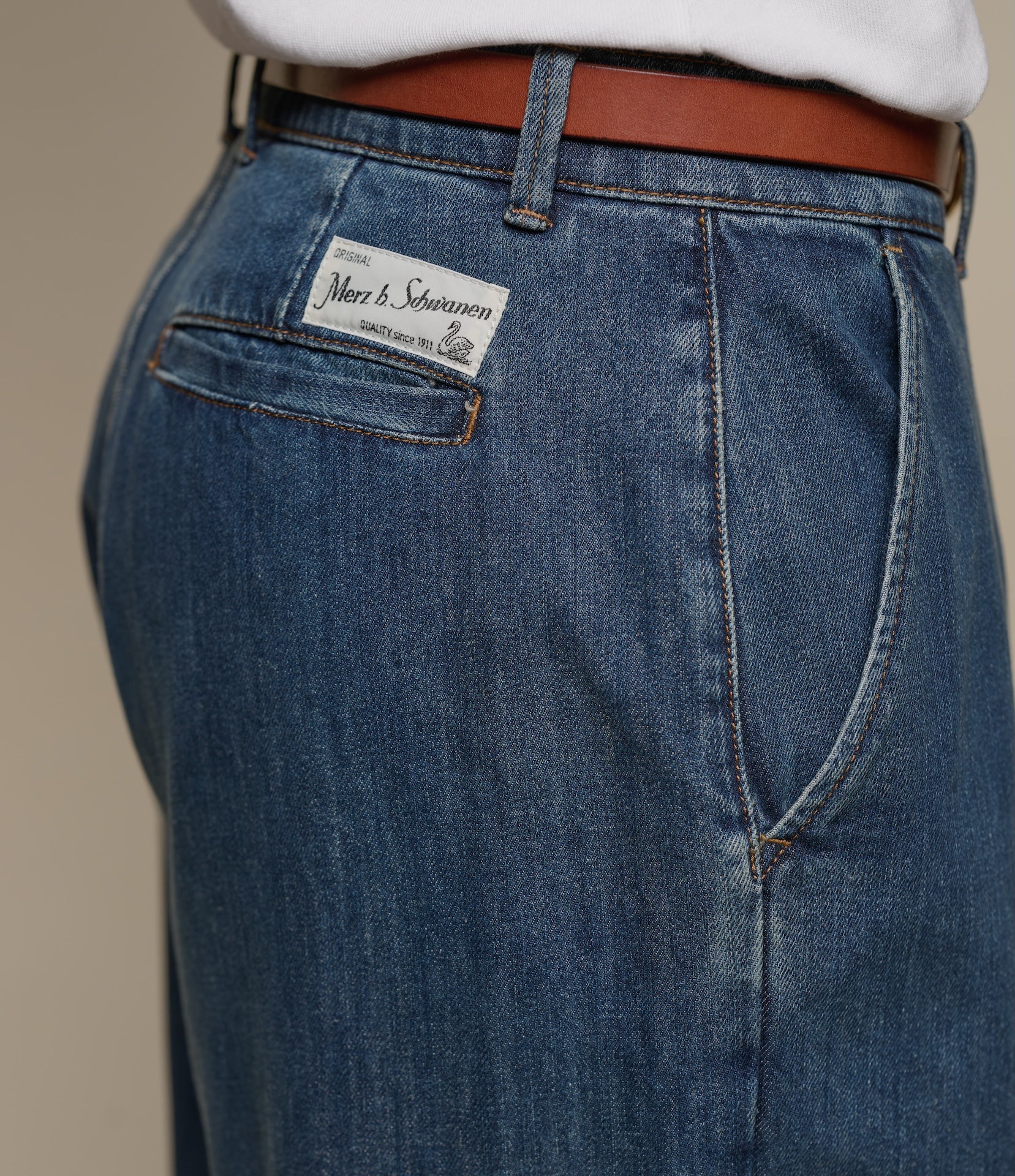 Close-up of blue jeans with a Merz b. Schwanen brand label on the back pocket against a beige background