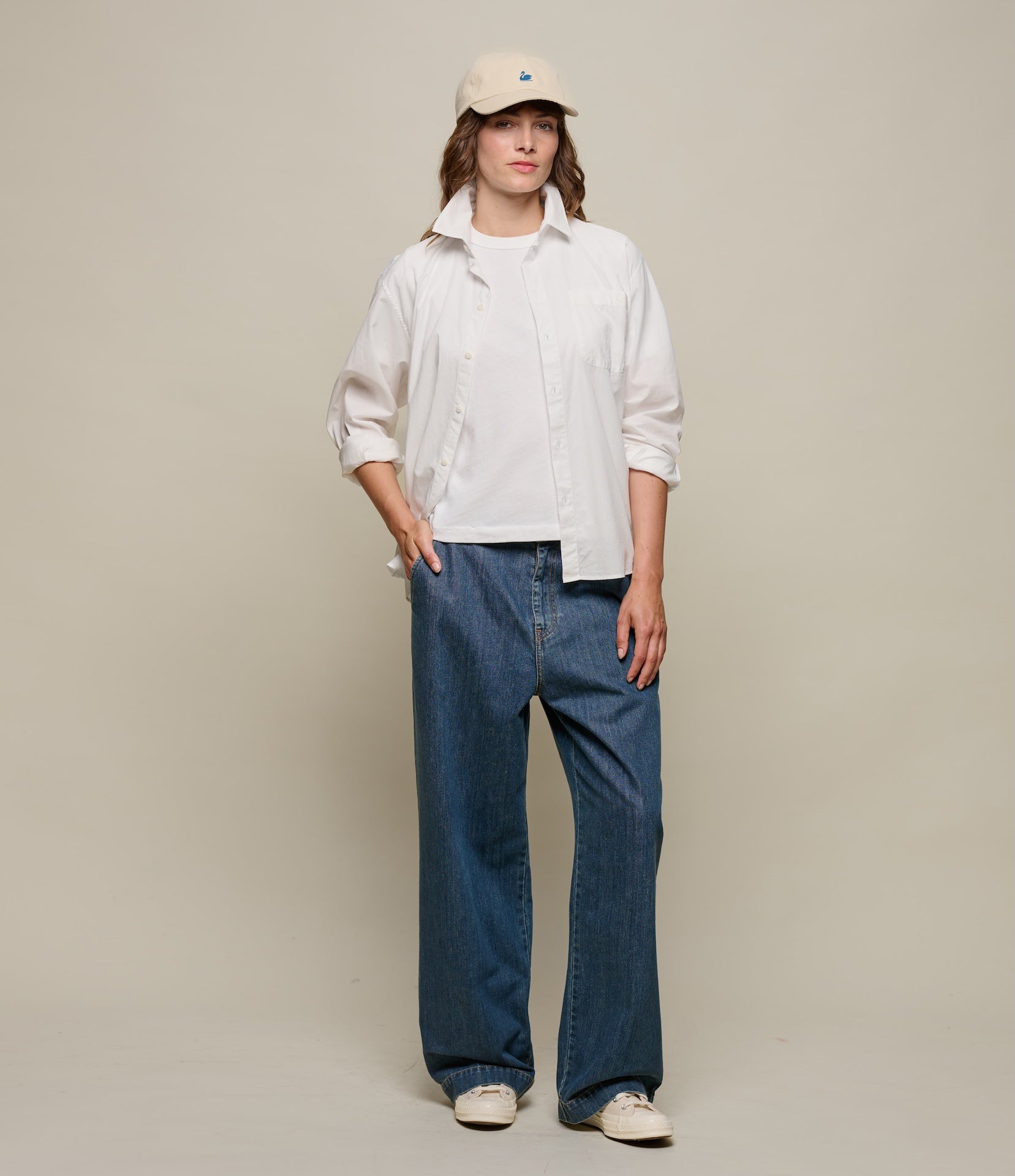 Person wearing a white shirt, blue jeans, and a beige cap on a plain background