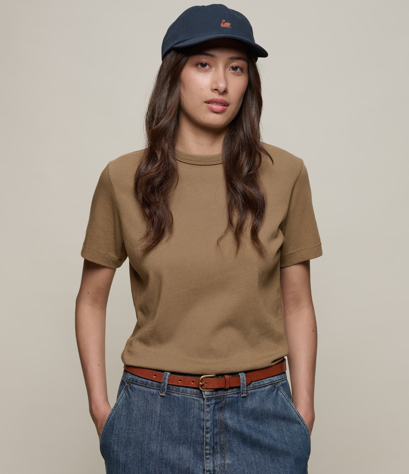 Person wearing a brown authentic jersey t-shirt, blue jeans, and a navy cap on a beige background