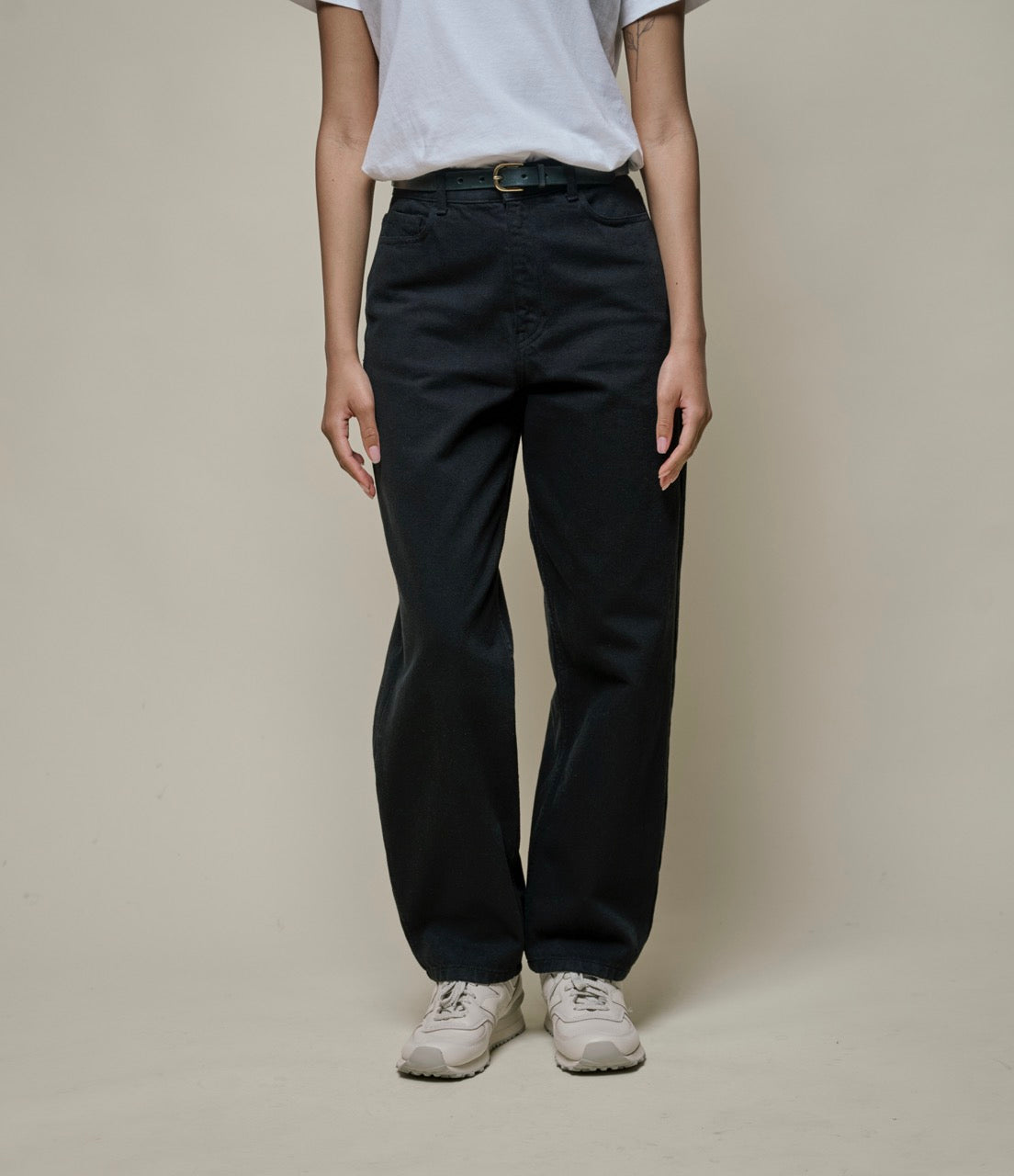 A person standing against a plain background, wearing black ankle-length 5-pocket-pants and white shoes.