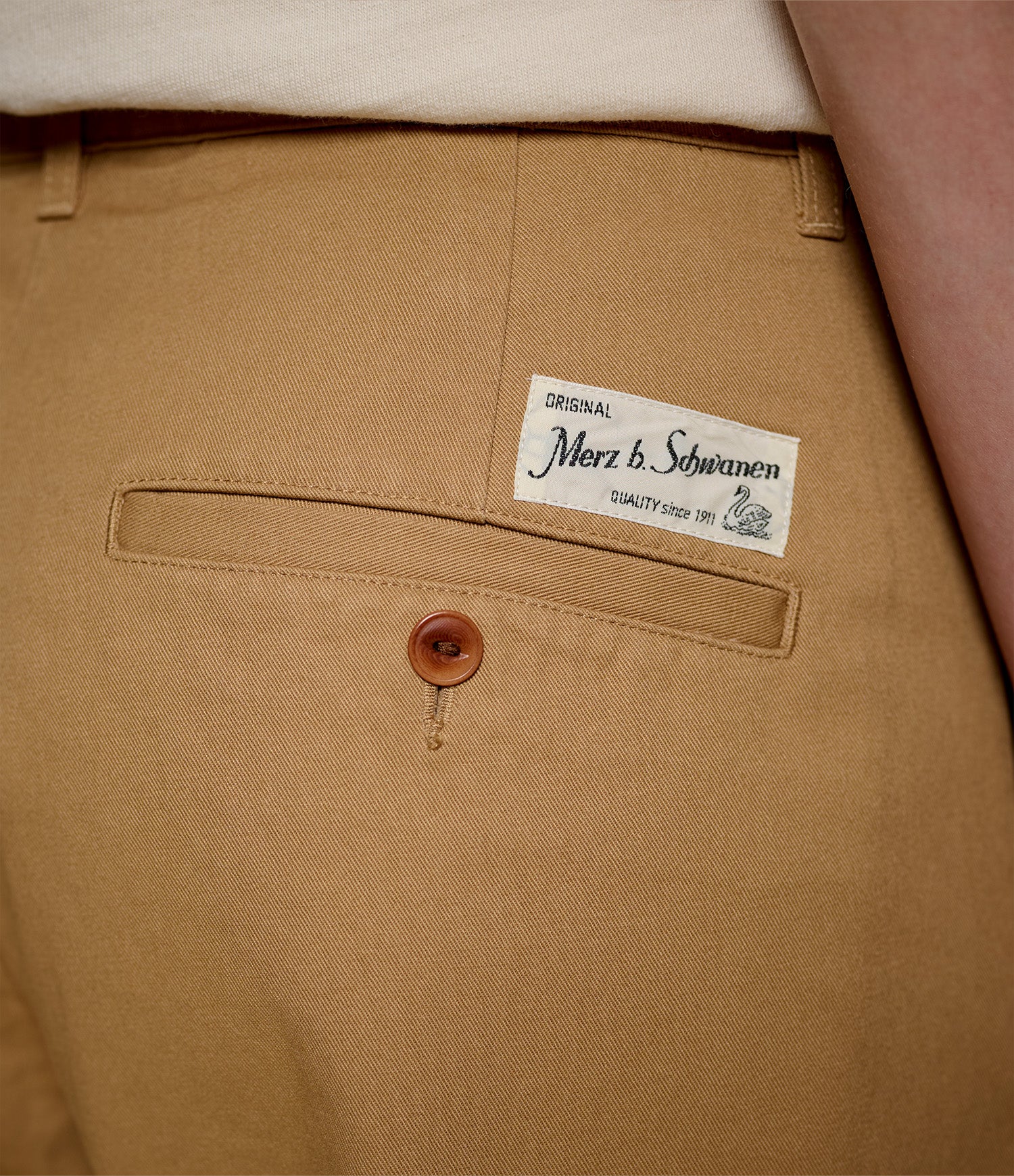 Close-up of tan chinos showing the back pocket with a brown button and a white brand label reading 'Original Merz b. Schwanen'.