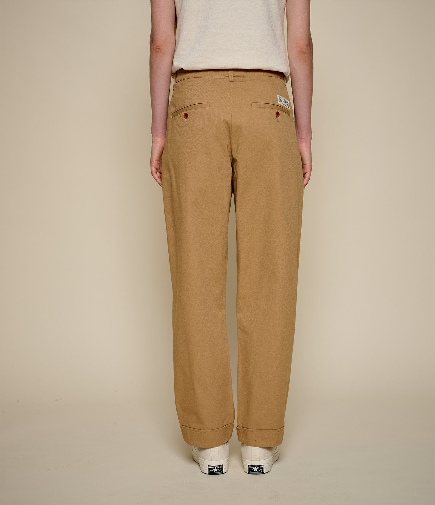 A person shown from the back wearing tan straight-leg pants, a light beige top, and white sneakers against a plain beige studio background.