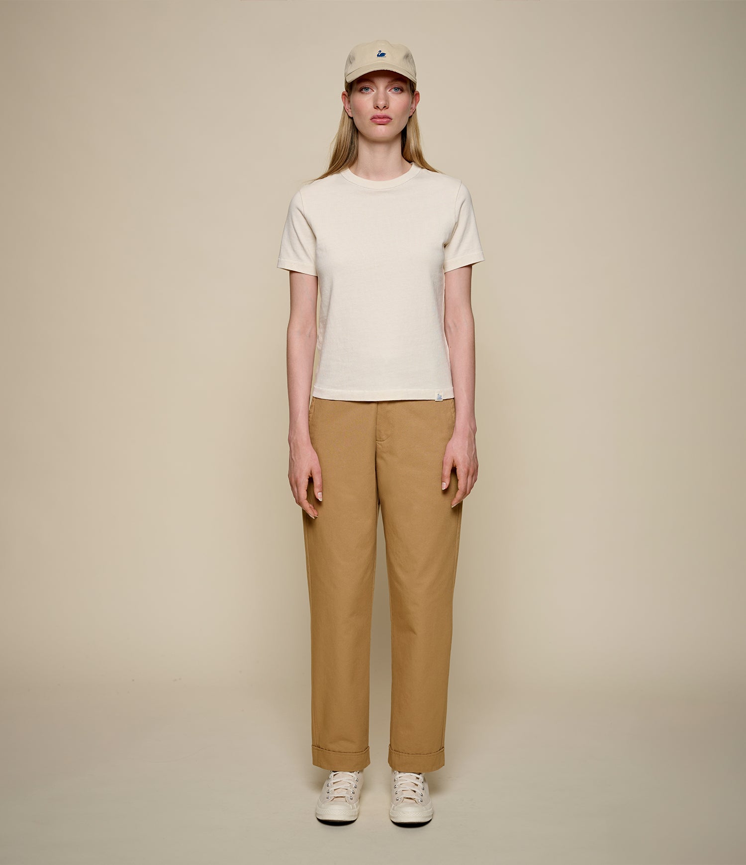 A model stands against a solid beige studio background wearing a cream-colored short-sleeve t-shirt, tan straight-leg pants, a beige baseball cap, and white sneakers.