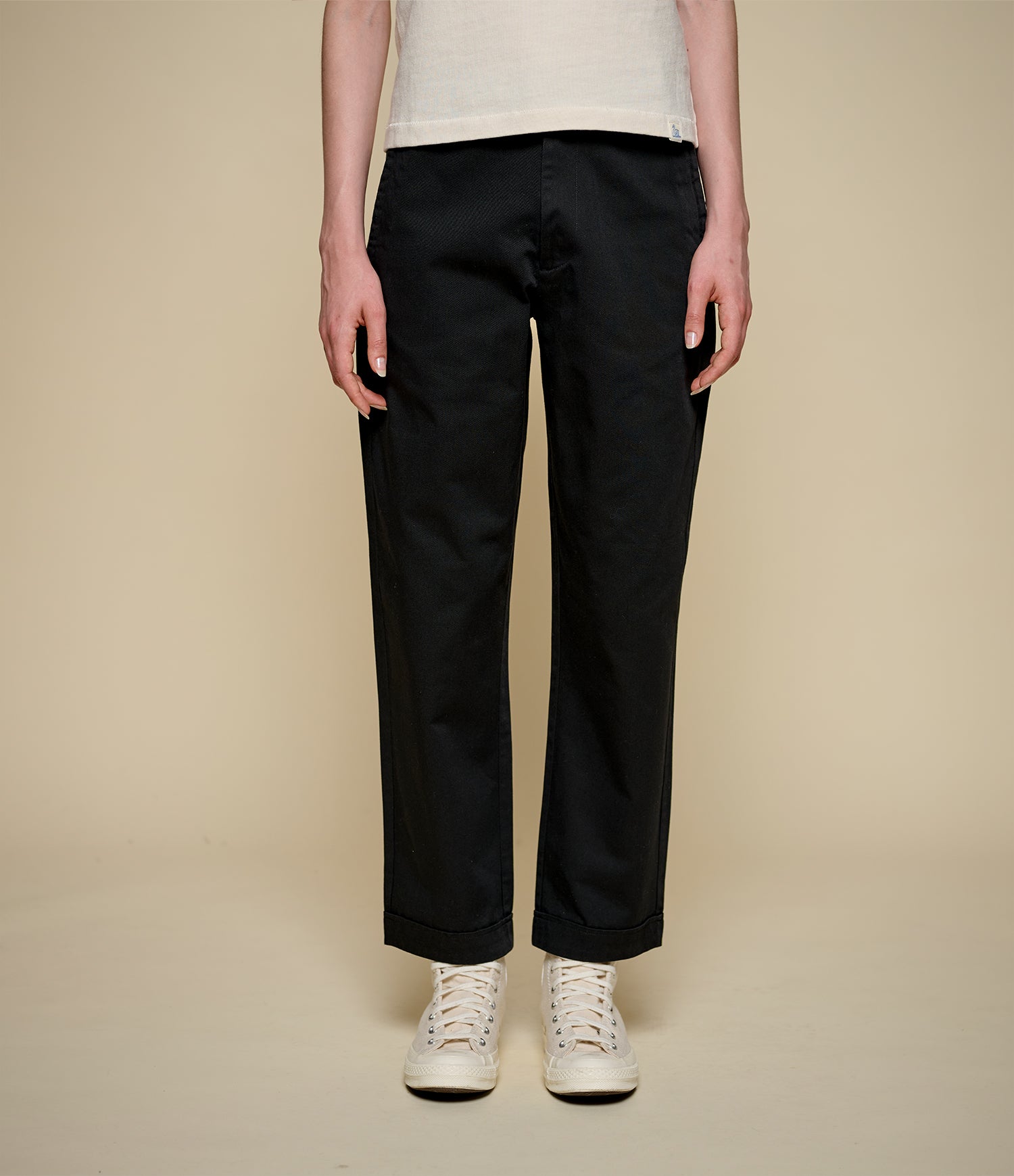 A model from the waist down wearing black straight-leg trousers, a cream t-shirt, and white sneakers, standing against a solid beige studio background.