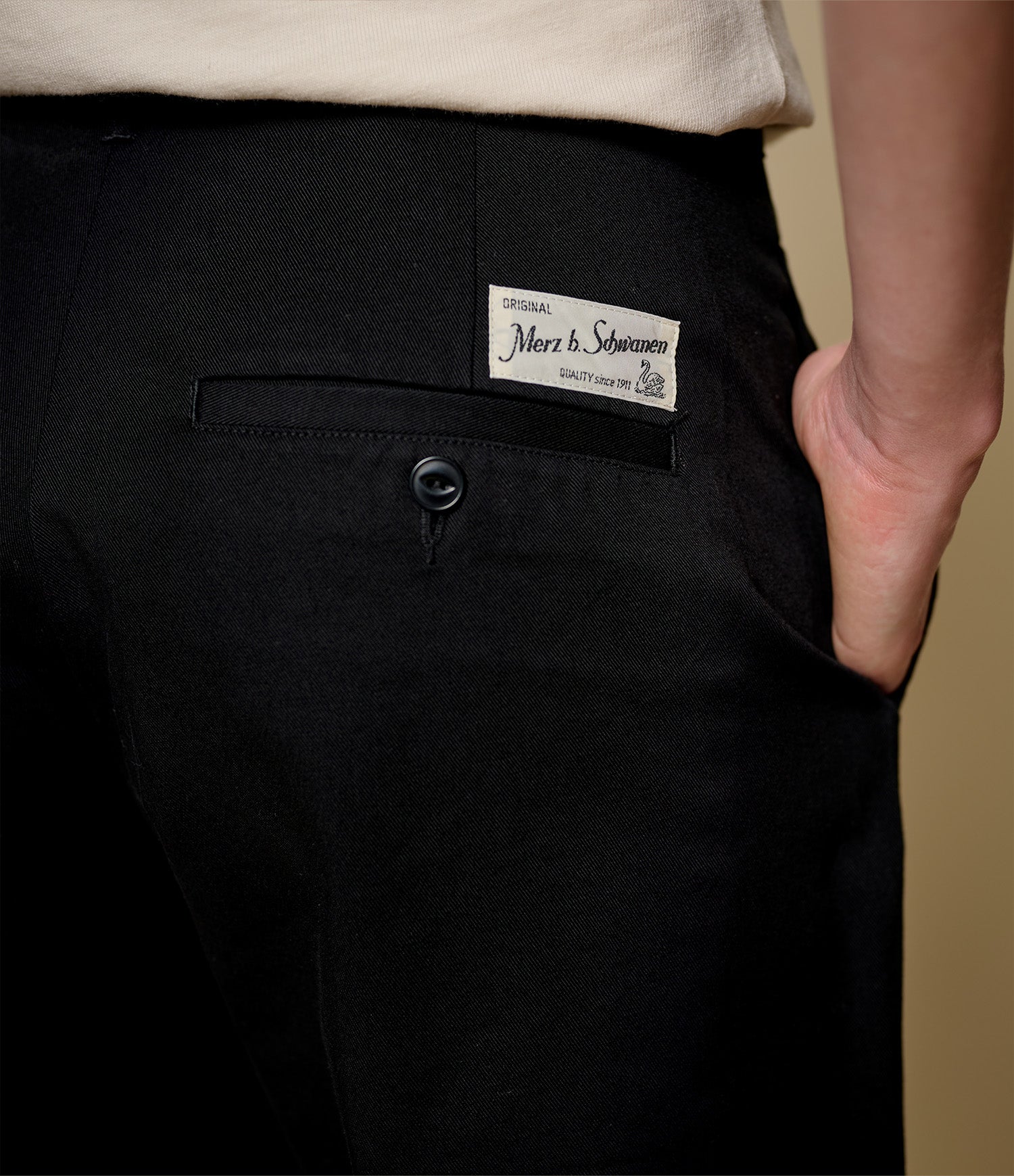 Close-up of a person wearing black trousers, showing the back pocket with a visible Merz b. Schwanen label and a hand in the pocket.