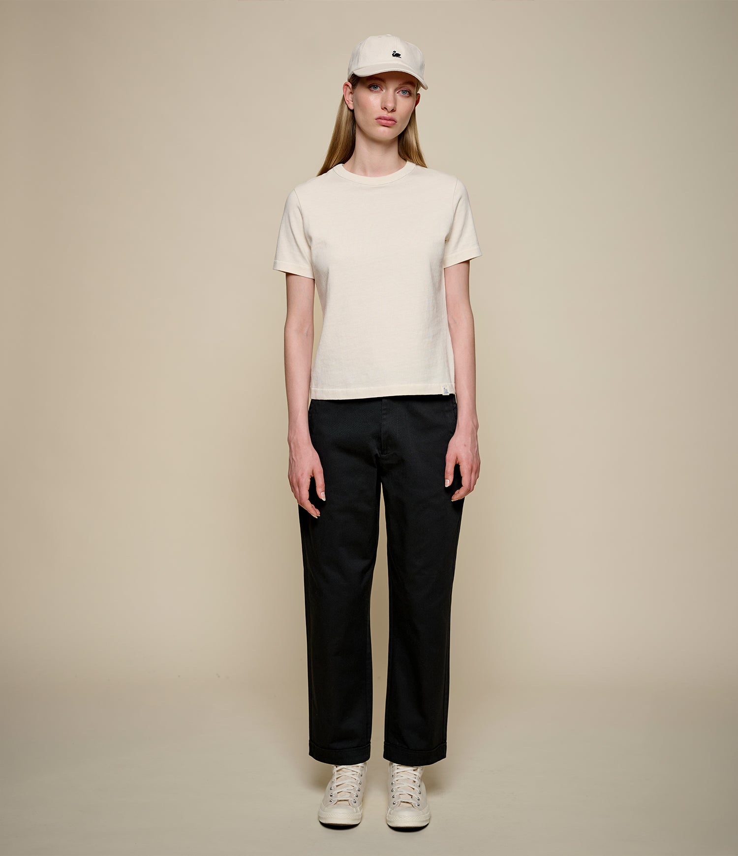 A model stands against a plain beige studio background wearing a cream-colored short-sleeve crewneck t-shirt, black wide-leg trousers, white sneakers, and a white baseball cap.