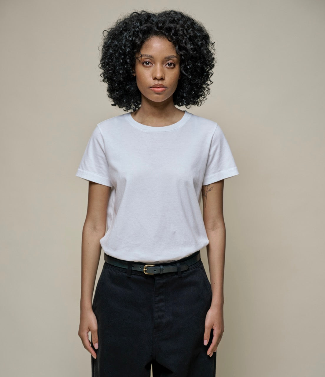 A female model wearing a plain white short-sleeve crewneck t-shirt and black pants, standing against a solid beige background.