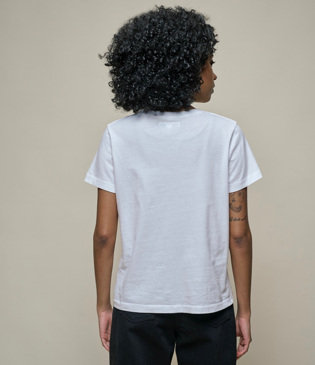A woman shown from the back wearing a plain white short-sleeve T-shirt and dark pants, standing against a plain beige studio background.