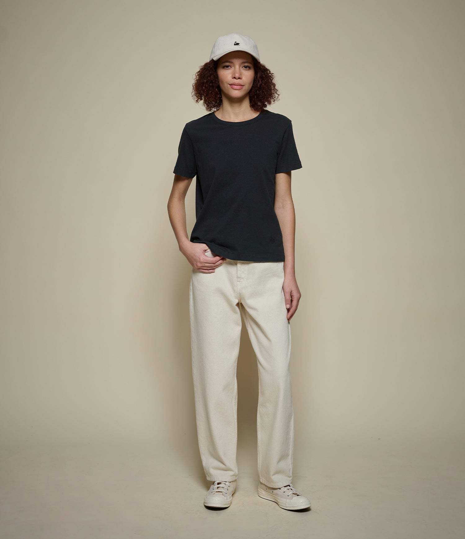Person wearing a black t-shirt, beige pants, and a white cap on a beige background