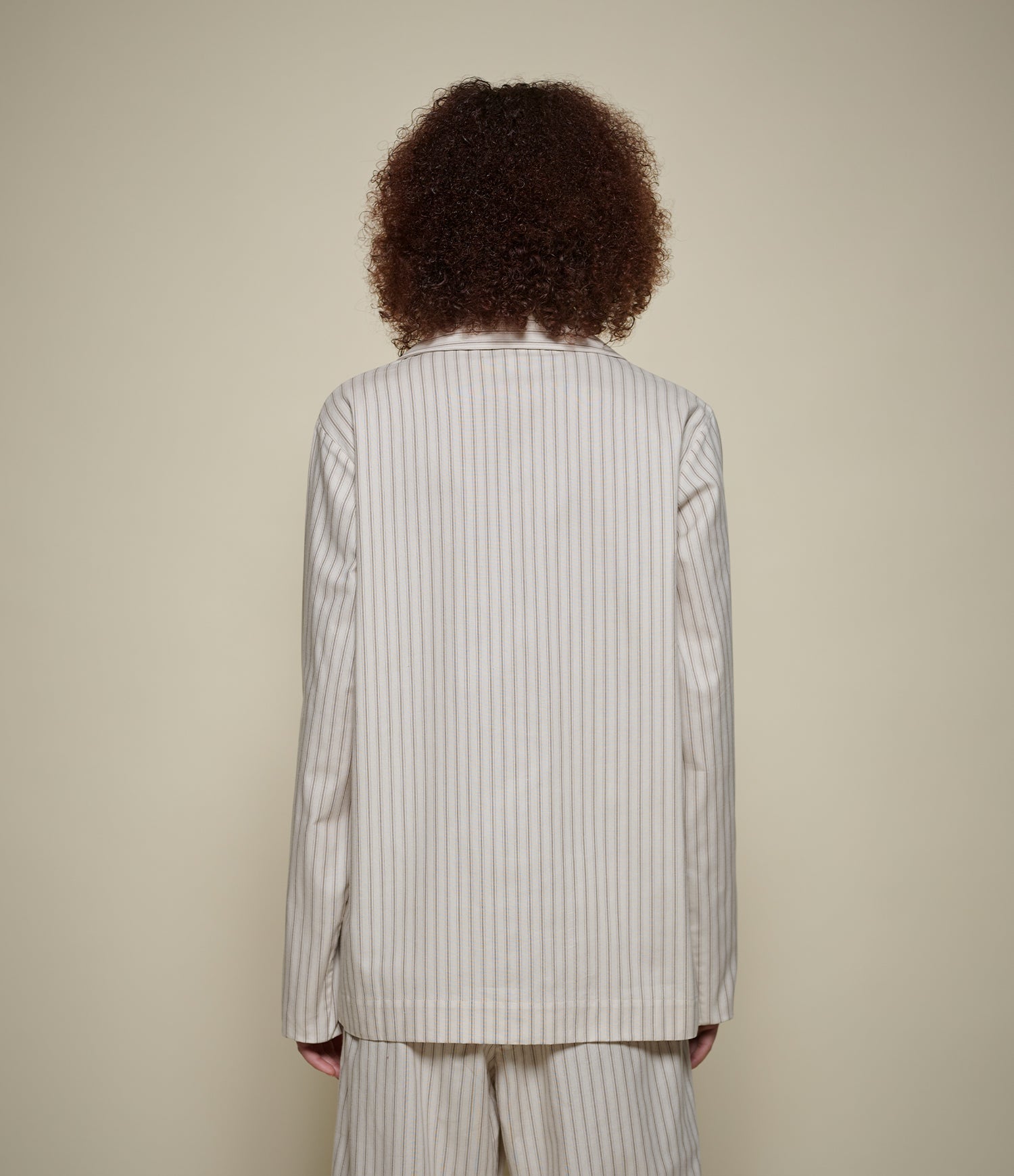 A person viewed from the back wearing a cream-colored pinstripe suit with thin dark vertical stripes, set against a solid light beige background.