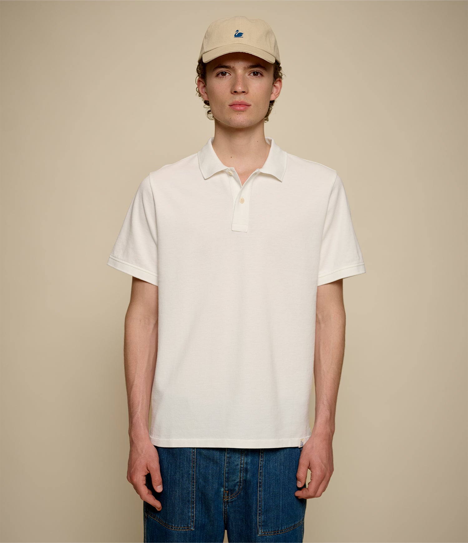Person wearing a beige cap and white pikée polo shirt against a beige background