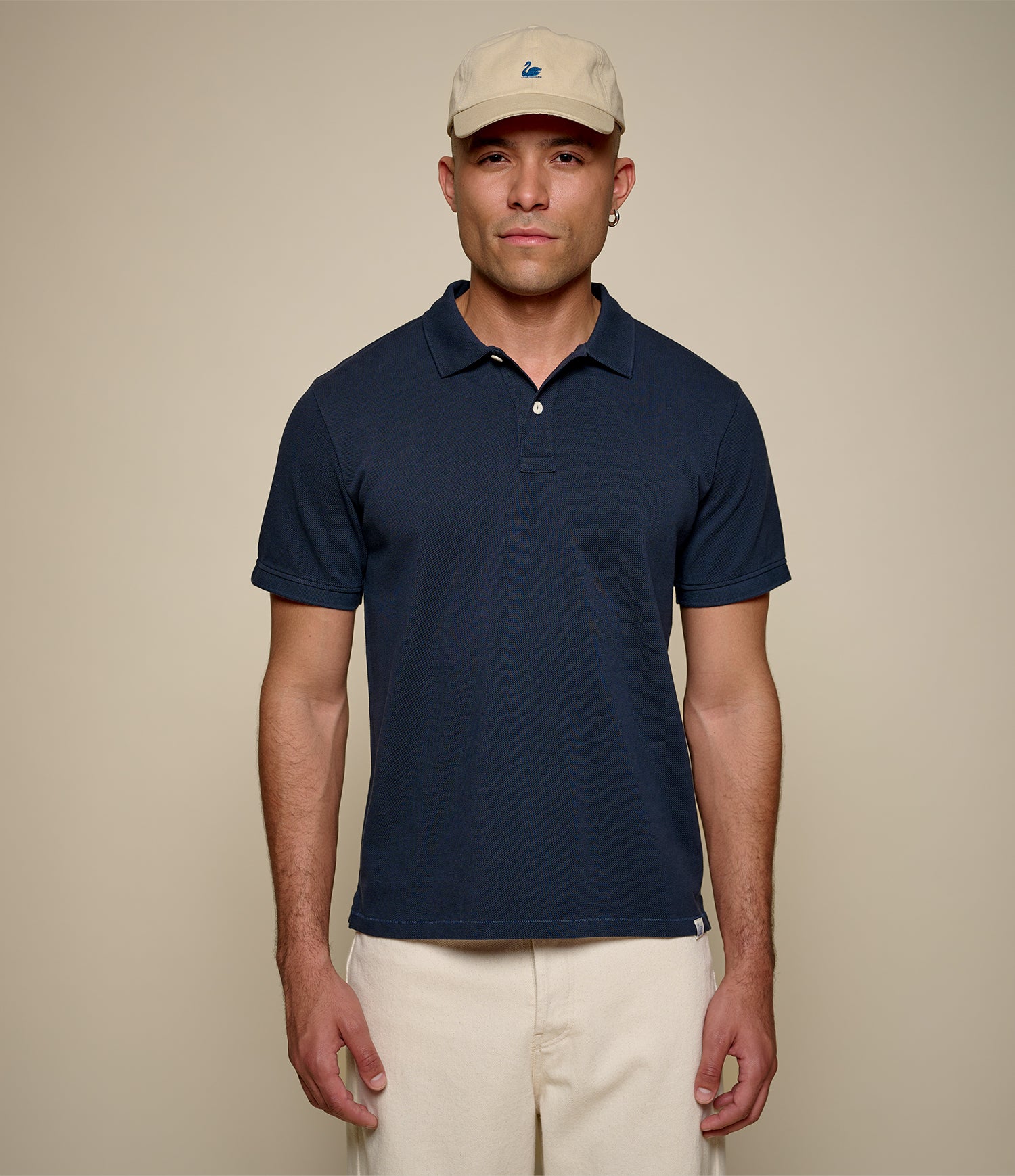 Man wearing a navy pikée polo shirt and beige cap against a beige background