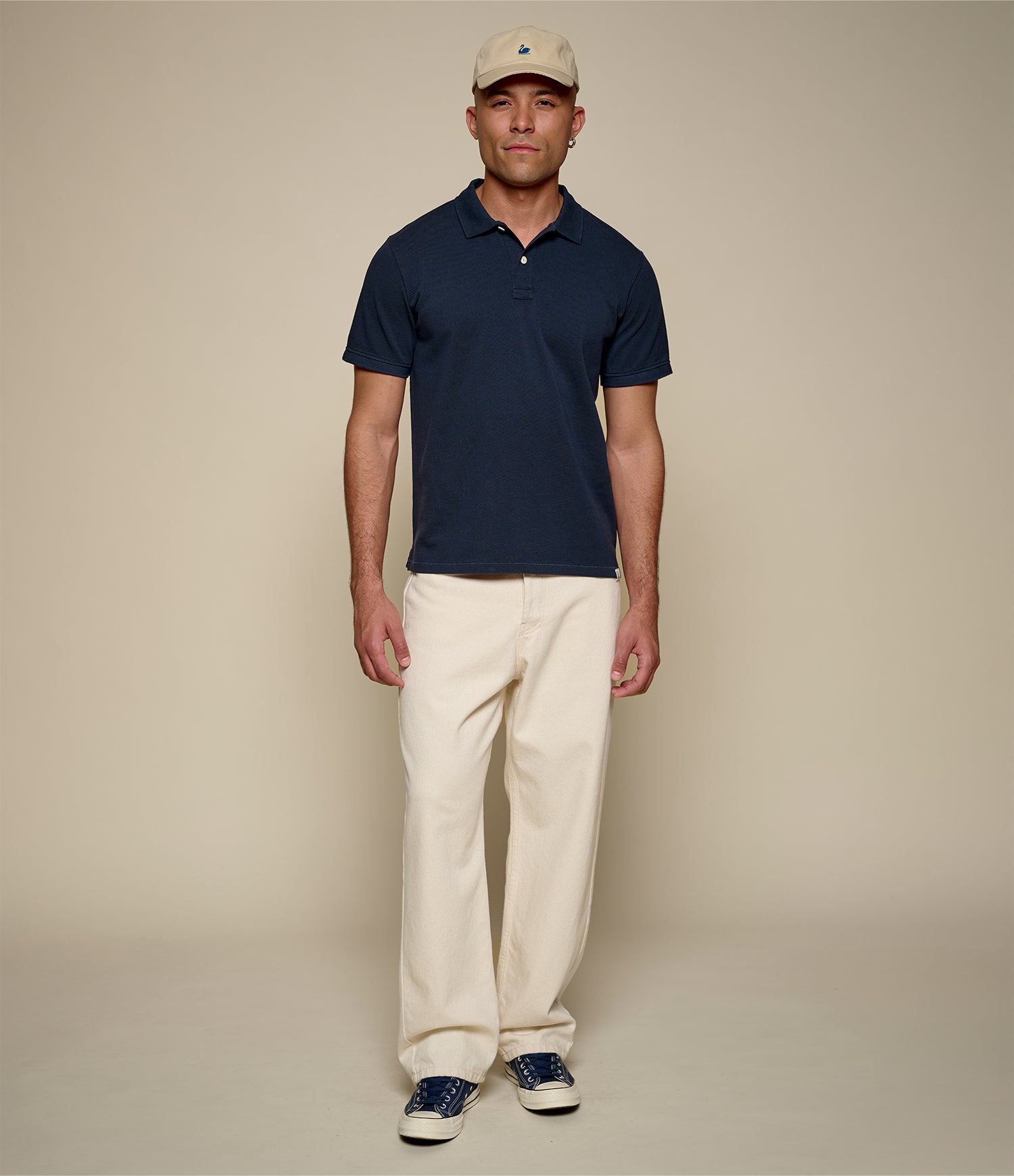 Man wearing a navy polo shirt, beige cap, and white pants against a beige background