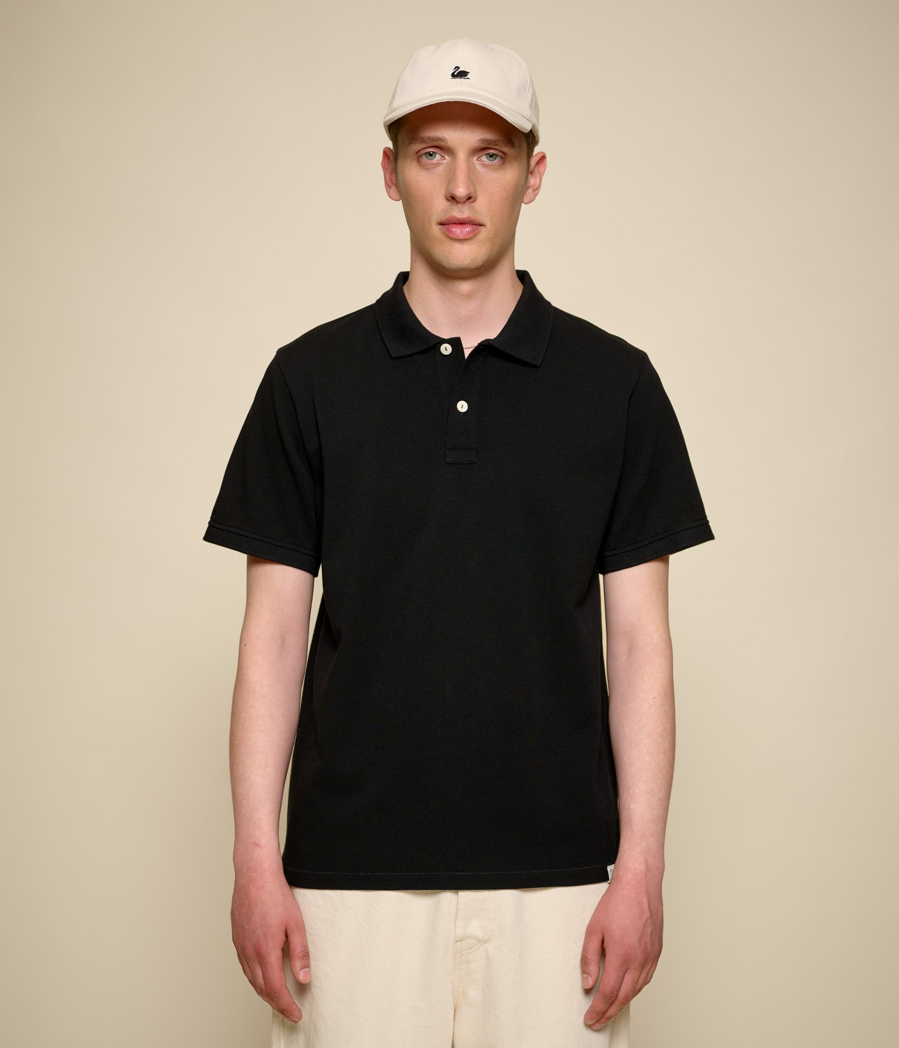 Man wearing a black pikée polo shirt and beige cap against a beige background
