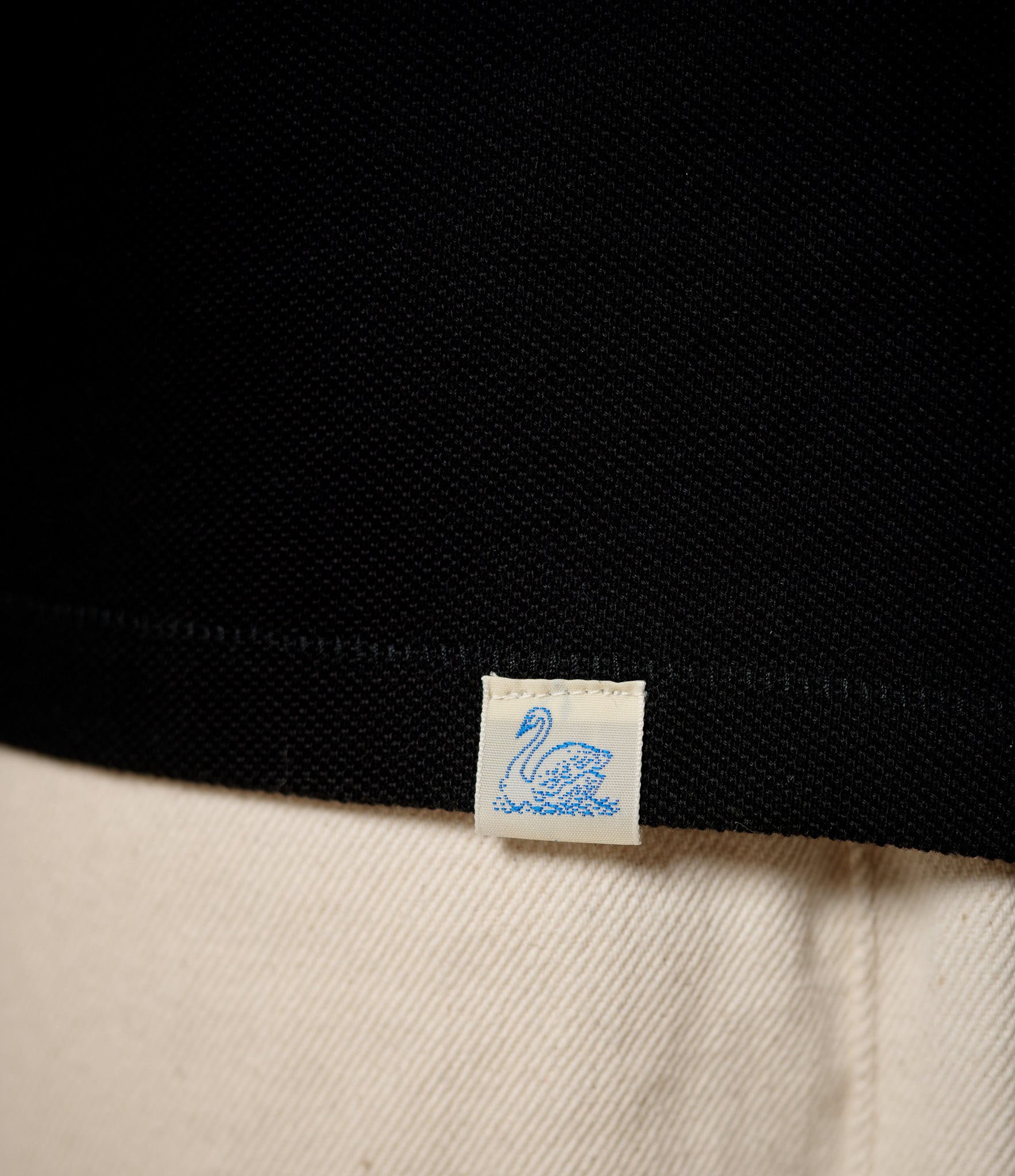 Close-up of a black pikée fabric with a blue label featuring a swan design on a beige background.