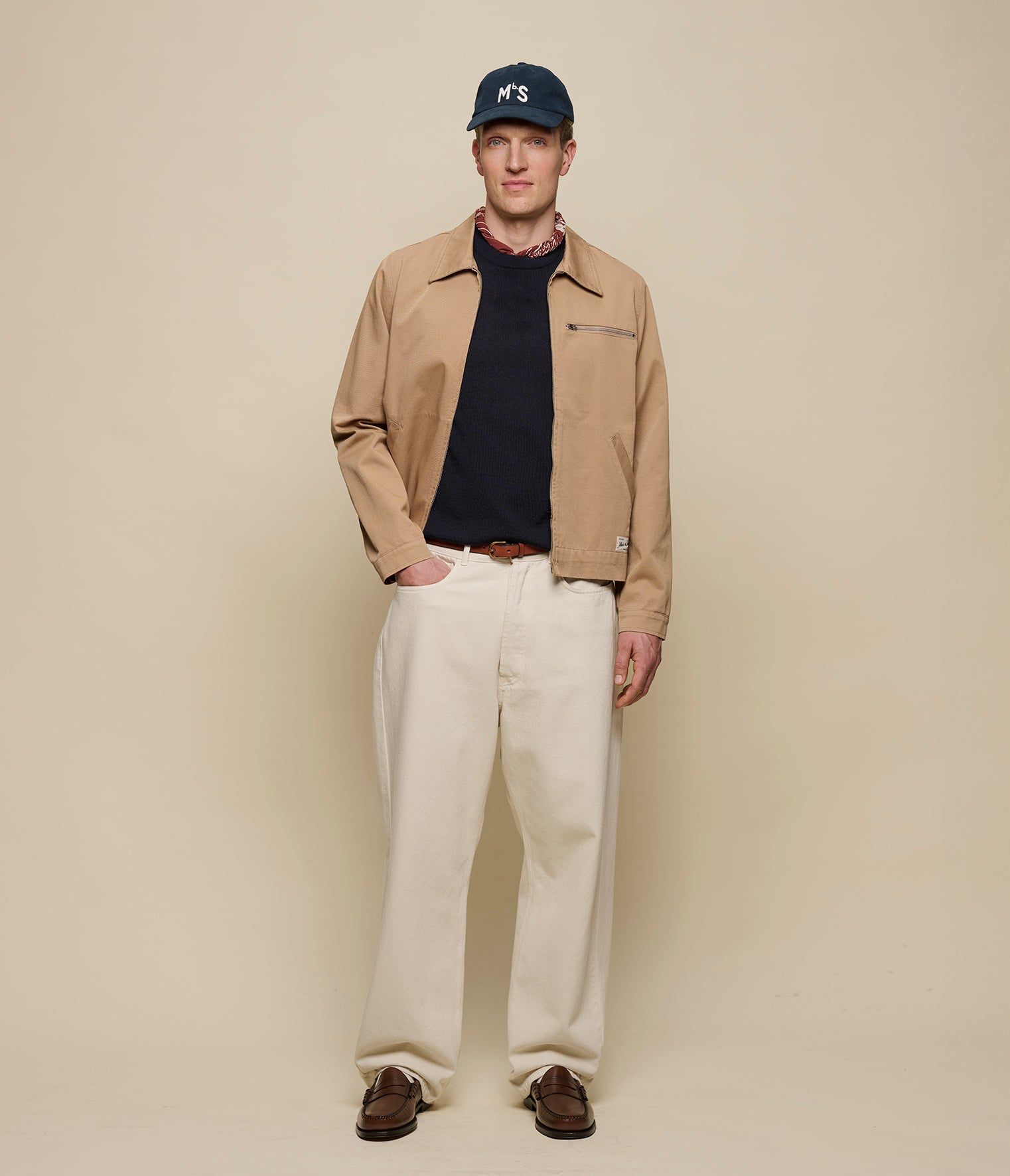 Man wearing a beige zip jacket, black shirt, white pants, and a navy cap against a beige background