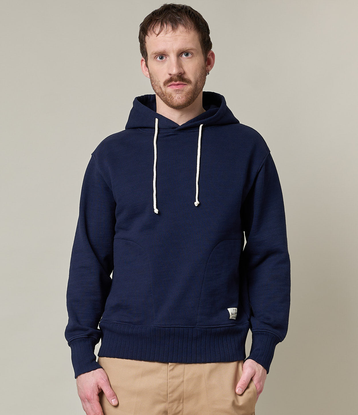 A male model wearing a navy blue hoodie with a hood and drawstrings, paired with beige pants.