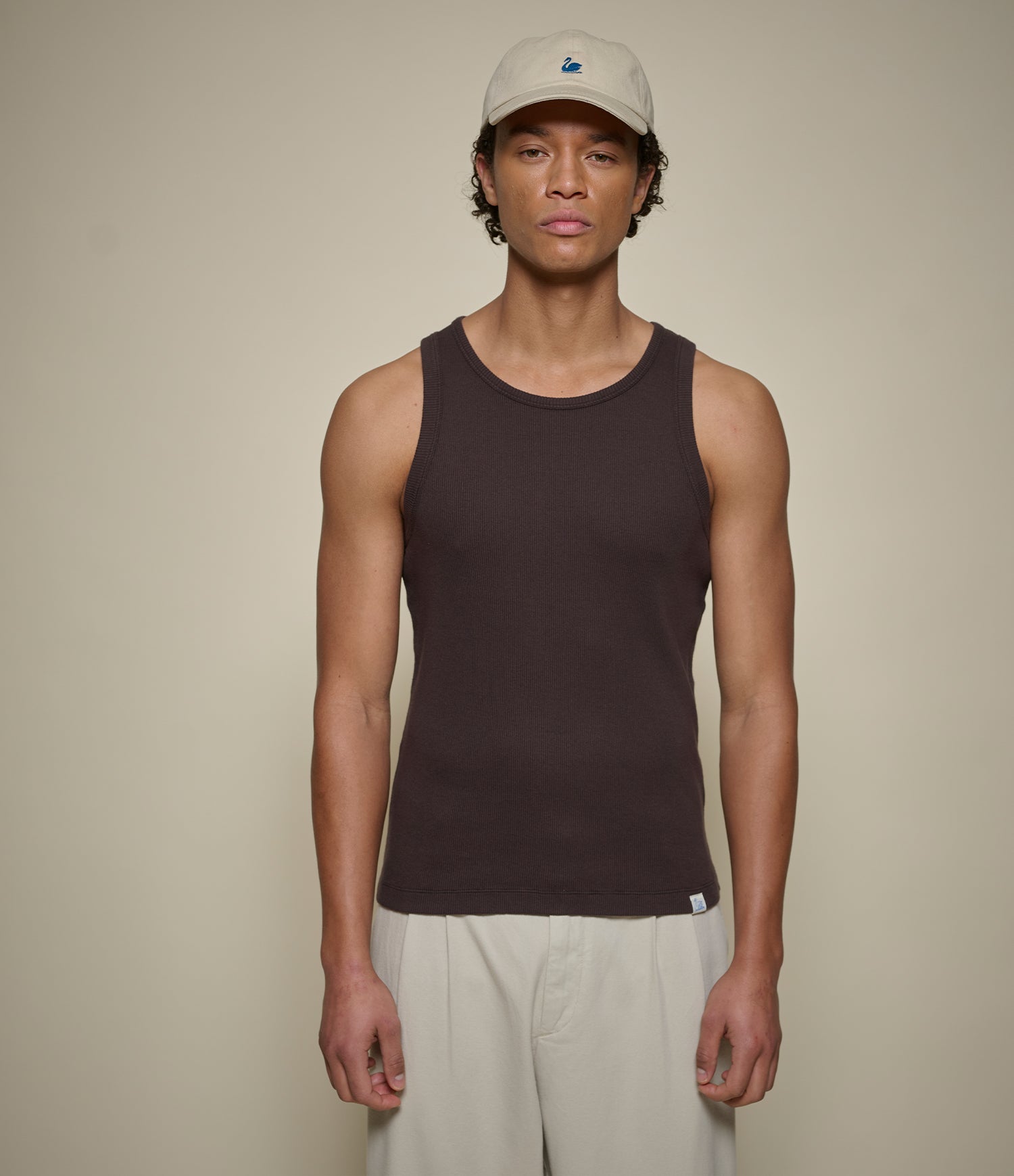 Man wearing a brown ribbed tank top and beige cap against a plain background