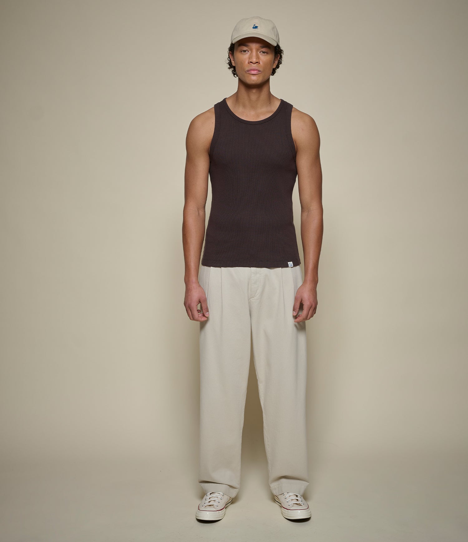 Person wearing a brown tank top, light pleated pants, and a cap on a beige background