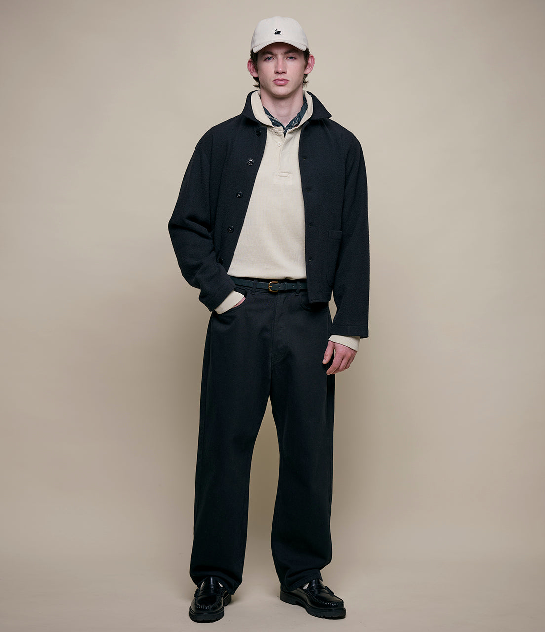 Man wearing a black jacket, white shirt, and black pants against a beige background