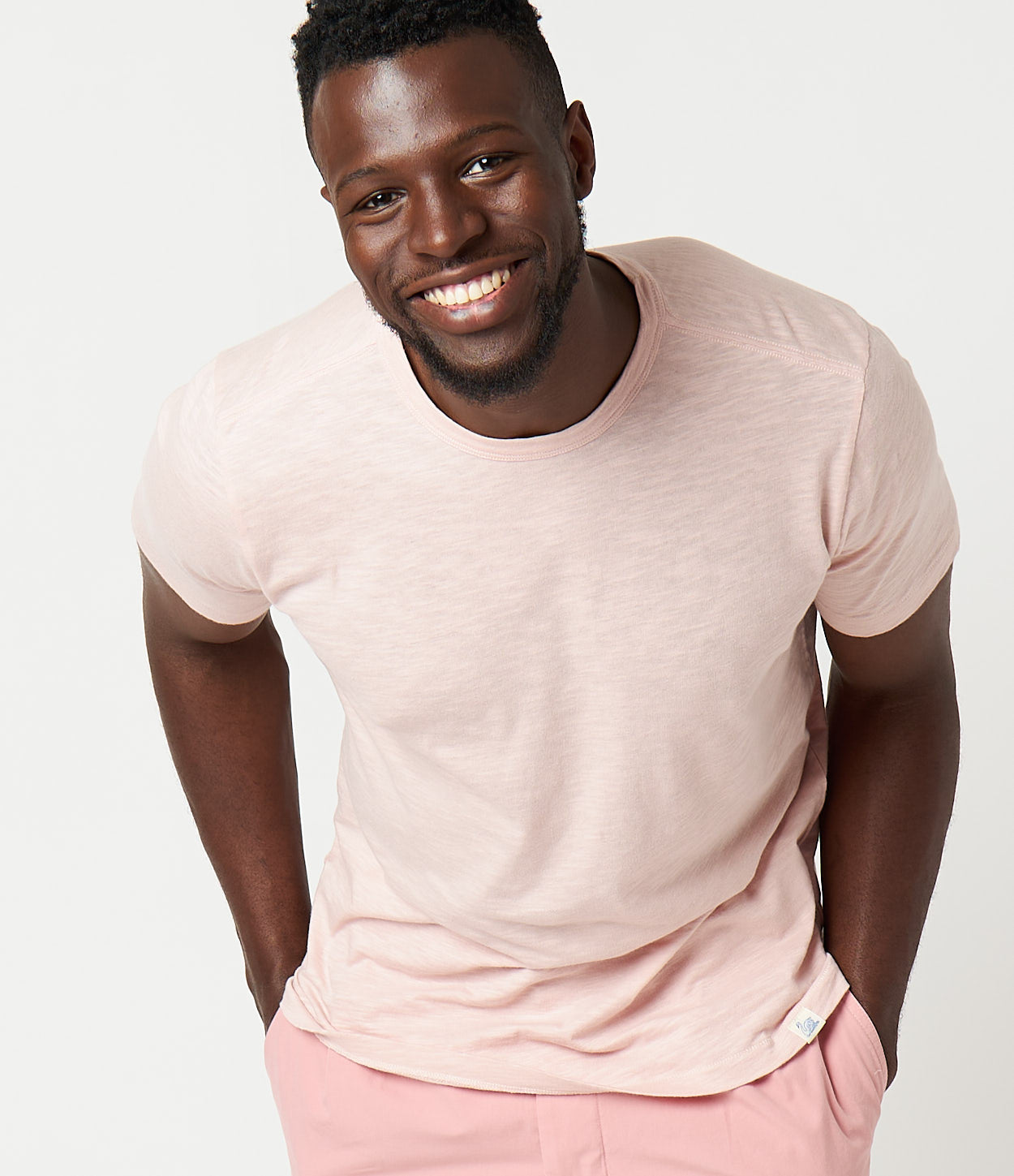 A man wearing a beige crew neck T-shirt and pink pants, standing against a white background.