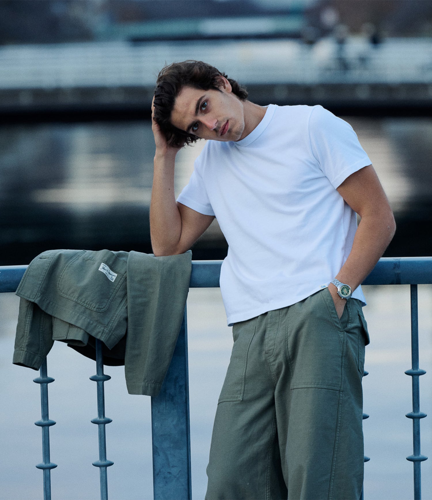 A man wearing a white crew-neck t-shirt and olive green cargo pants leans against a blue railing with water in the background. An olive green jacket is draped over the railing next to him.