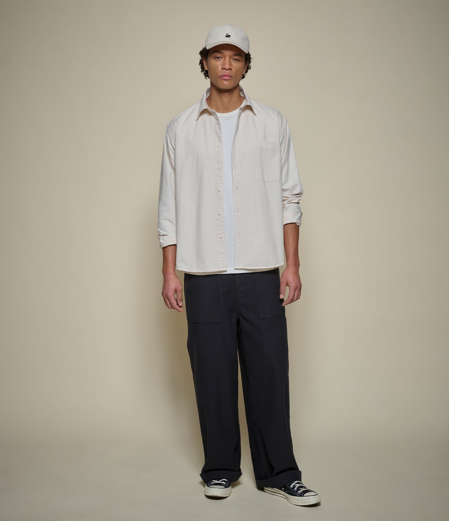 A man stands against a plain beige studio background wearing a light cream button-up shirt over a white tee, dark navy pants, black sneakers, and a white cap.