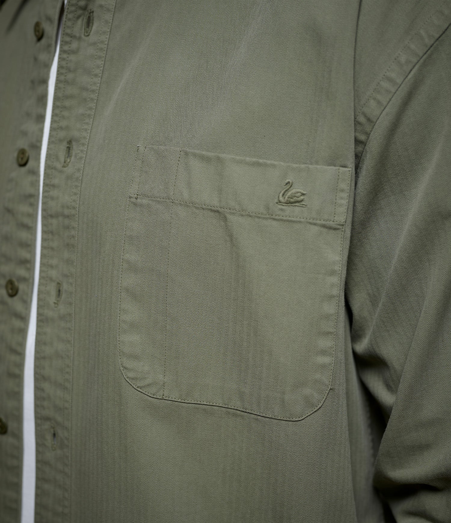 Close-up of an olive green button-up herringbone twill shirt featuring a chest pocket with a small embroidered Swan logo.