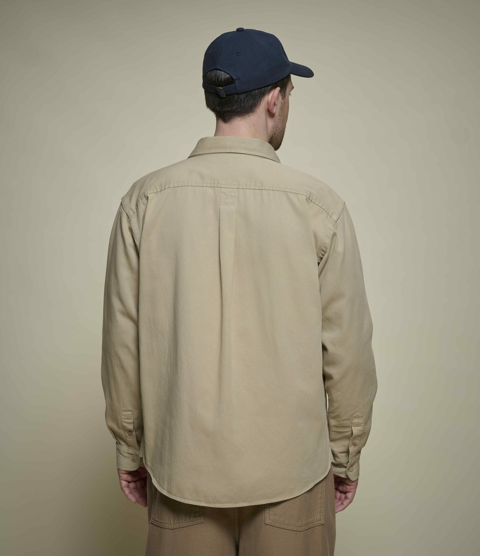 A man viewed from behind wearing a beige long-sleeve button-up shirt, khaki pants, and a navy blue baseball cap against a plain beige background.
