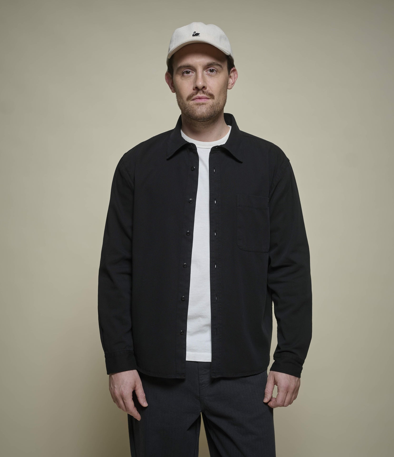 A man wearing a black button-up overshirt over a white t-shirt, dark pants, and a white baseball cap, standing against a solid beige background.