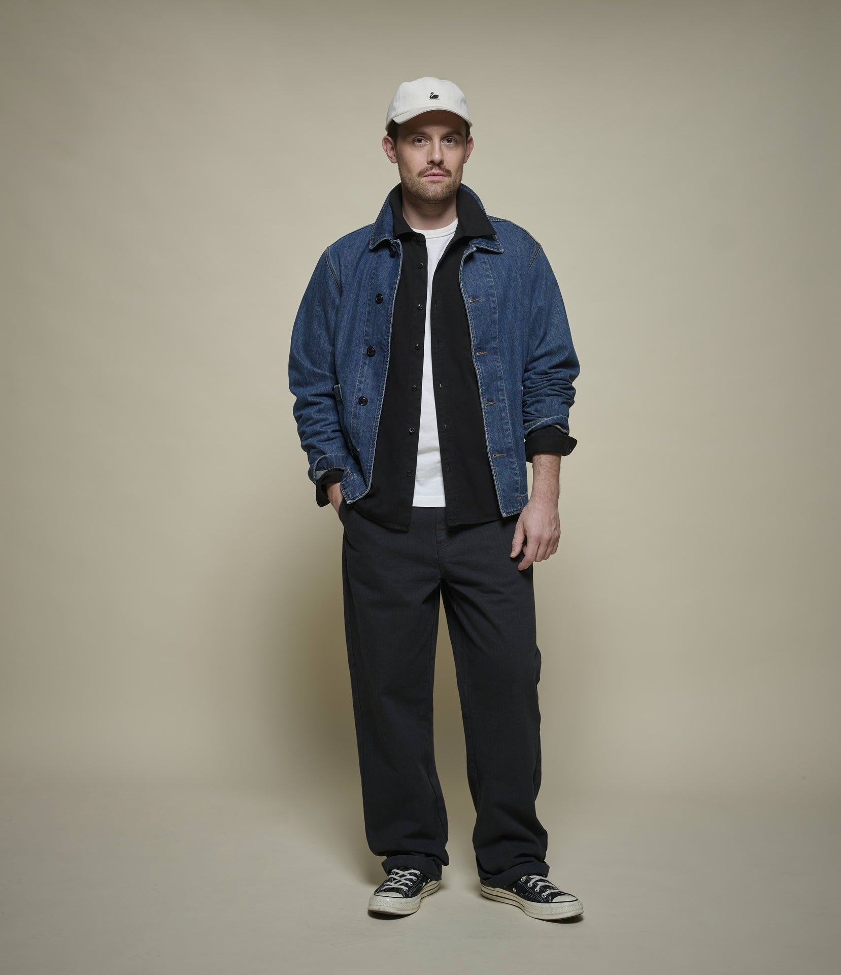 A man stands against a plain beige studio background wearing a blue denim jacket over a black shirt and white T-shirt, dark pants, black sneakers, and a white baseball cap.