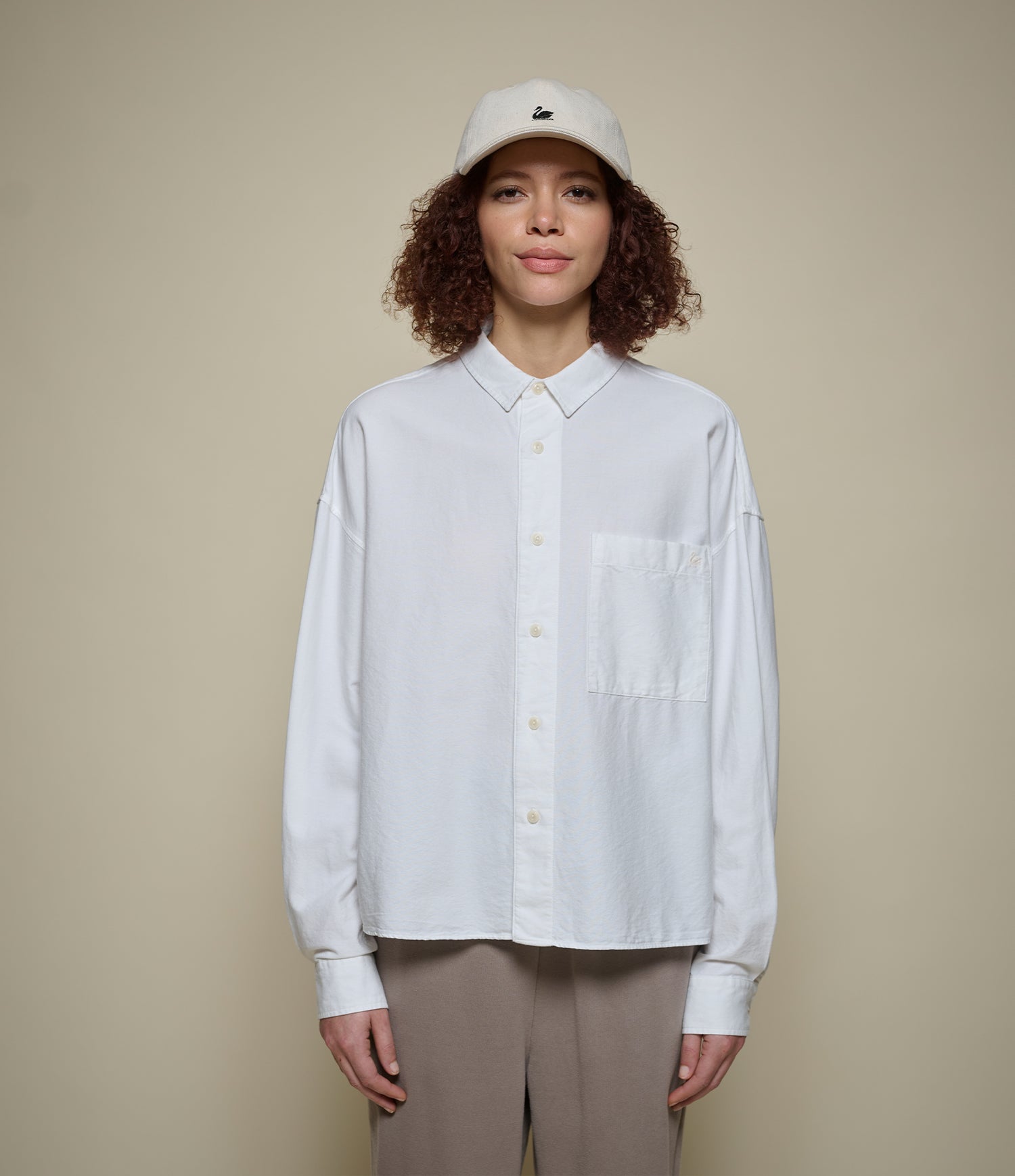 Person wearing a white  cropped oxford shirt and beige cap against a beige background