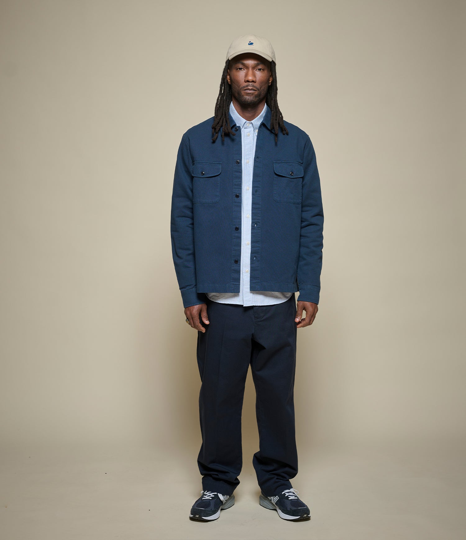 Man wearing a blue jacket, white shirt, dark pants, and a cap on a beige background