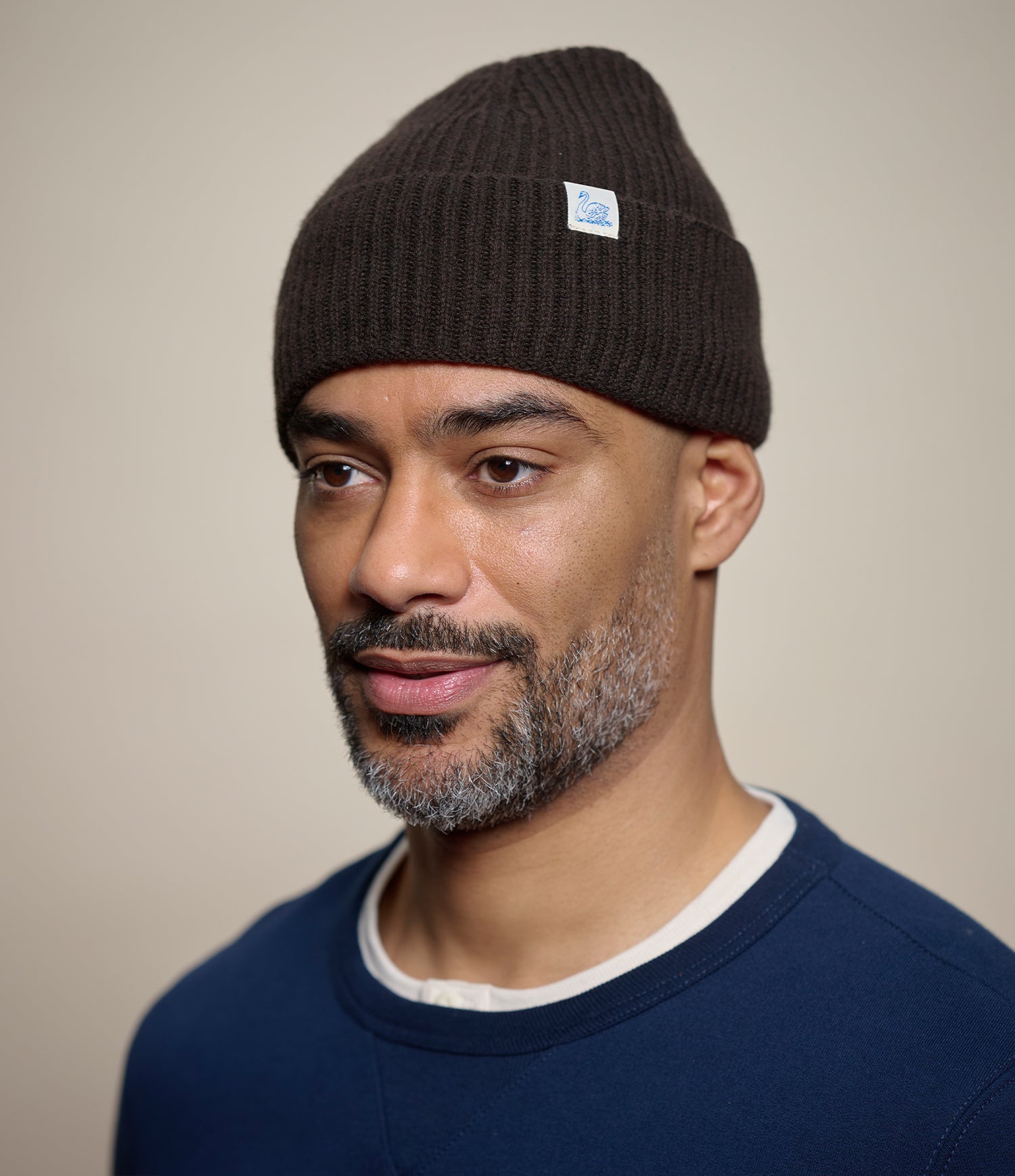 A man wearing a dark brown ribbed knit beanie with a small white and blue logo patch on the side, paired with a navy blue crewneck sweater. The background is a solid, warm neutral beige.