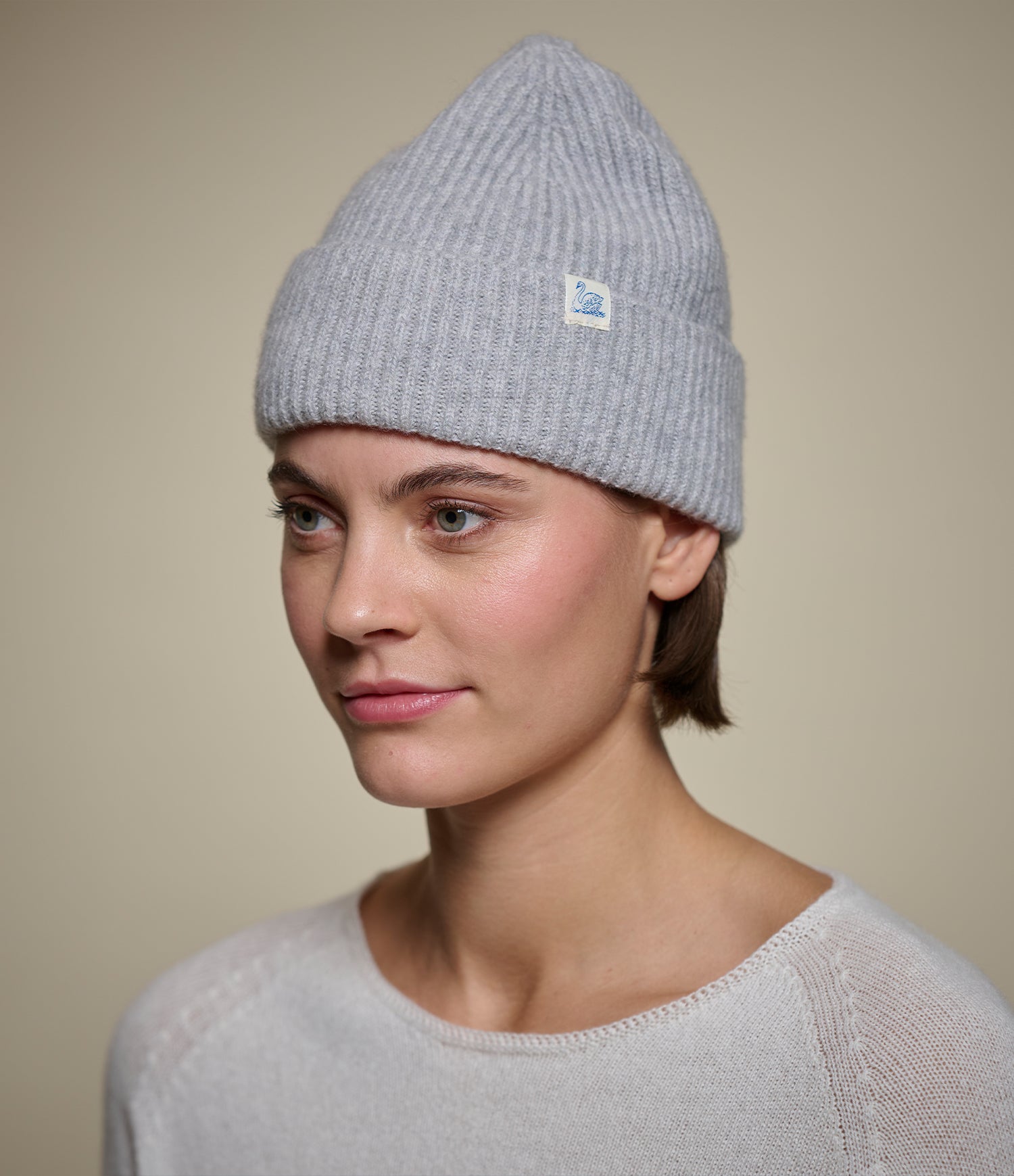A woman wearing a light gray ribbed knit beanie with a small white Swan brand tag on the side, photographed against a plain beige background.