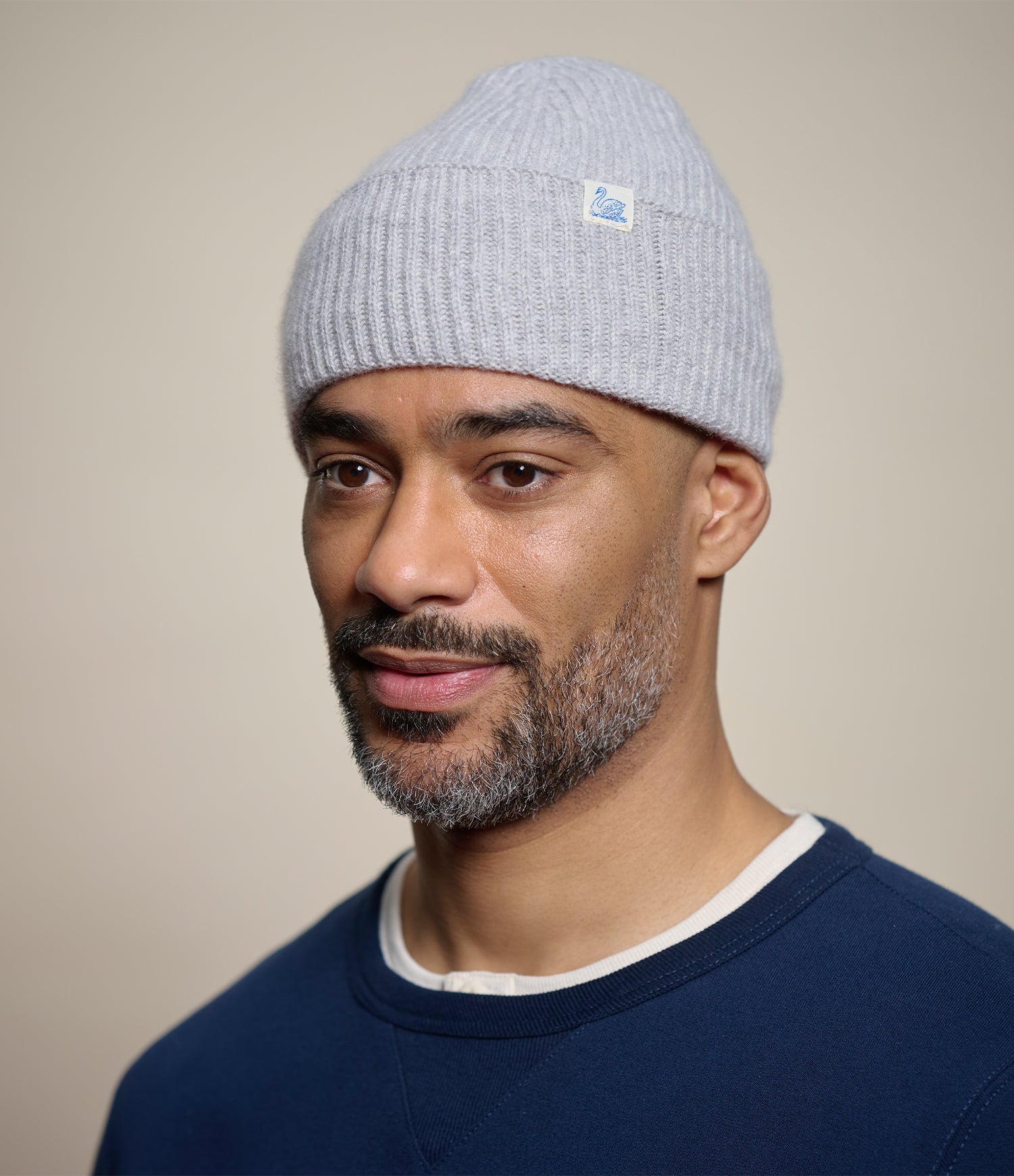 A man wearing a light gray ribbed knit beanie with a small swan brand patch on the side, paired with a navy crewneck sweater against a plain beige background.
