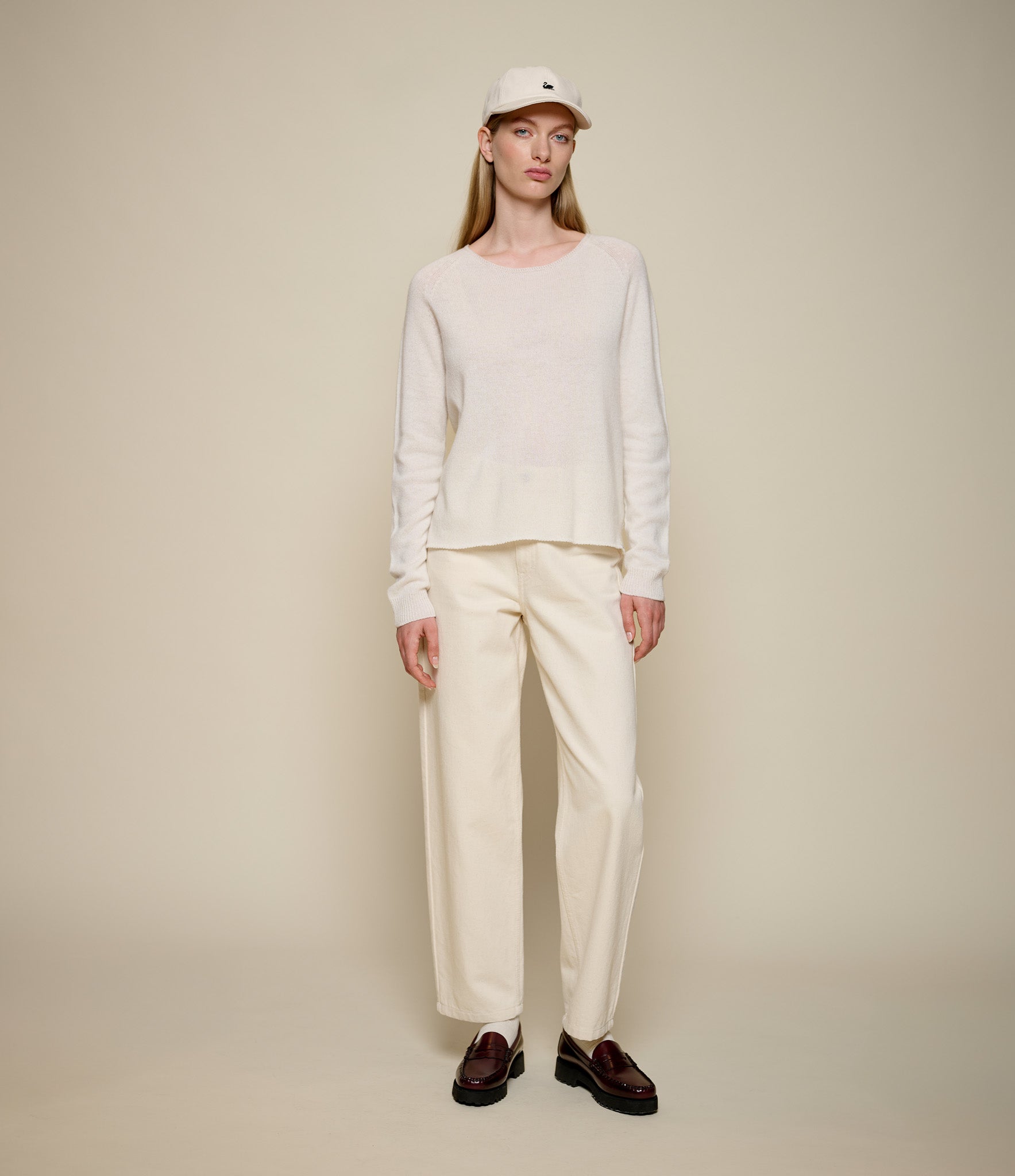 Person wearing a beige sweater and pants with a beige cap on a plain background