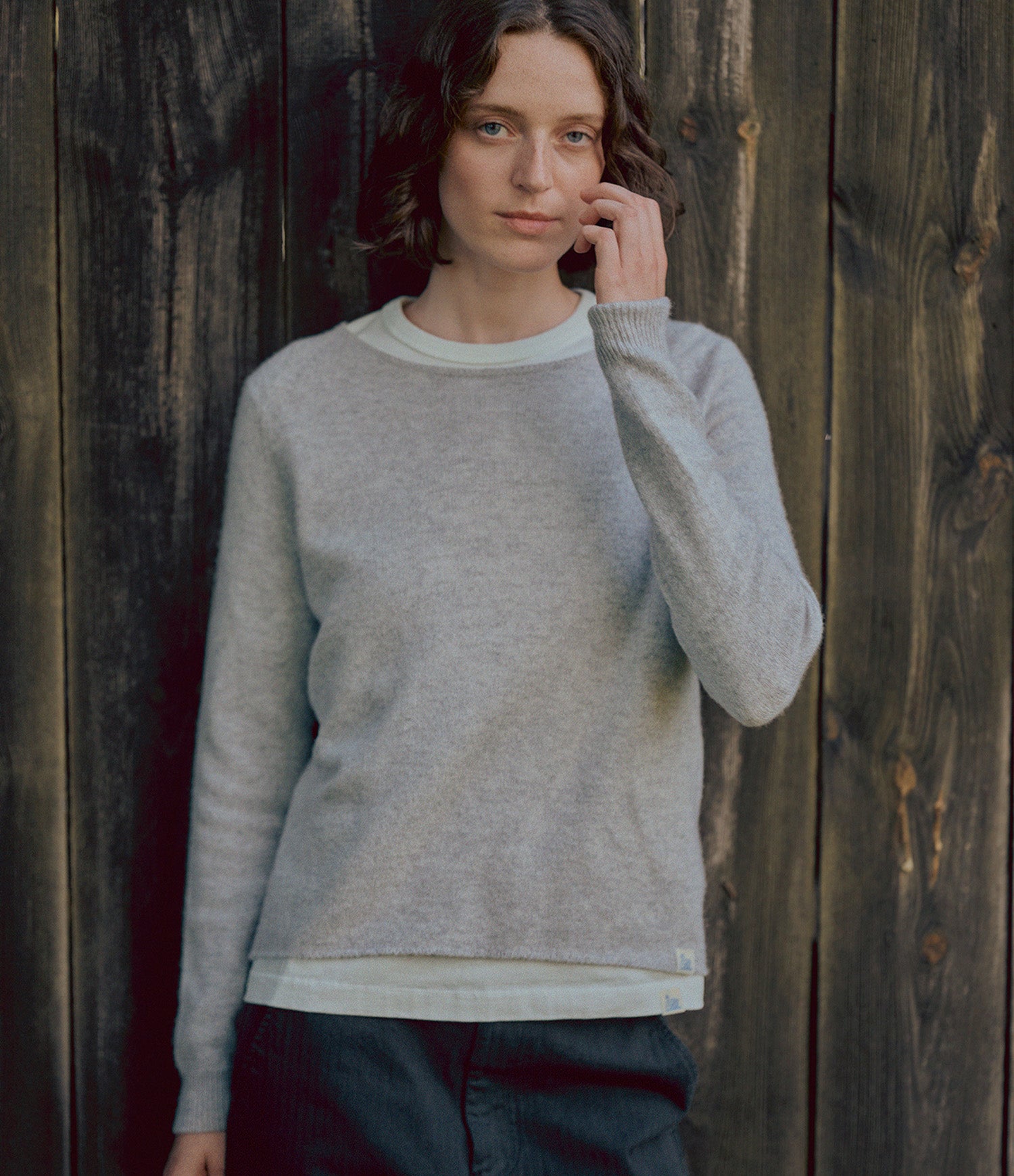 A woman wearing a light gray knit crewneck sweater over a white undershirt, standing against a dark rustic wooden plank wall.