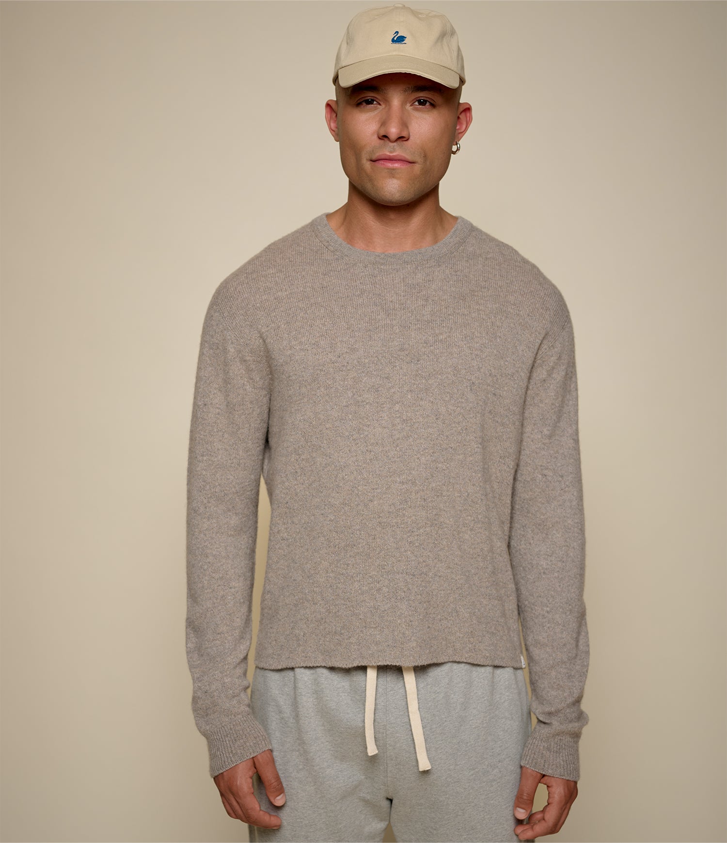 Man wearing a beige cap, gray sweater, and light gray pants against a beige background