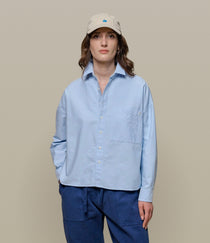 Person wearing a light blue cropped oxford shirt and dark blue pants with a beige cap on a beige background