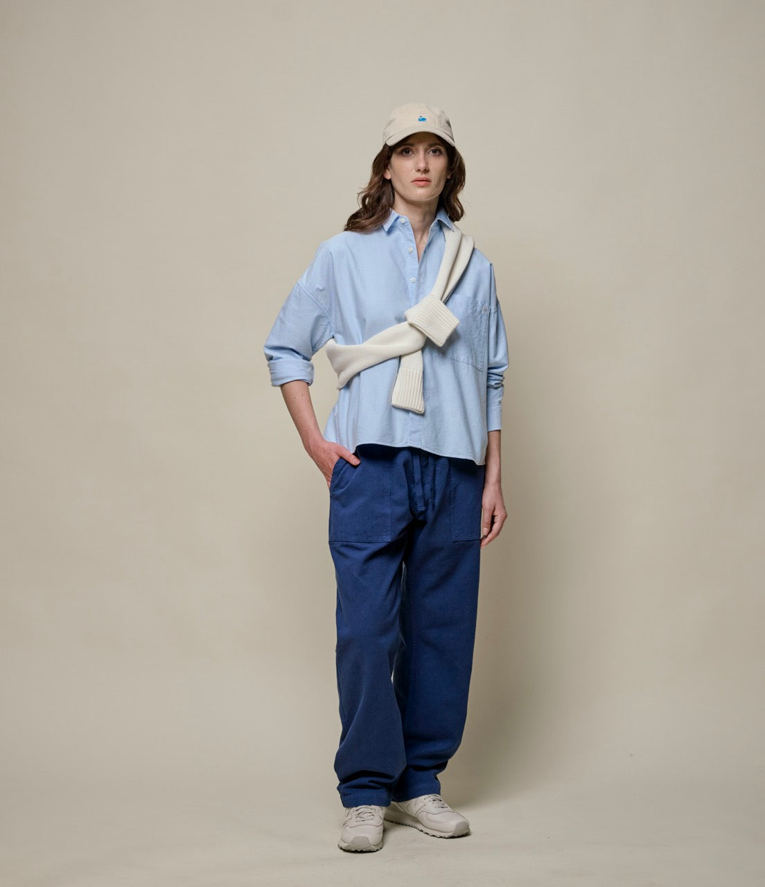 women wearing a blue cropped ocxford shirt, blue twill pants and a beige cap with a beuge sweater wrapped around her shirt.
