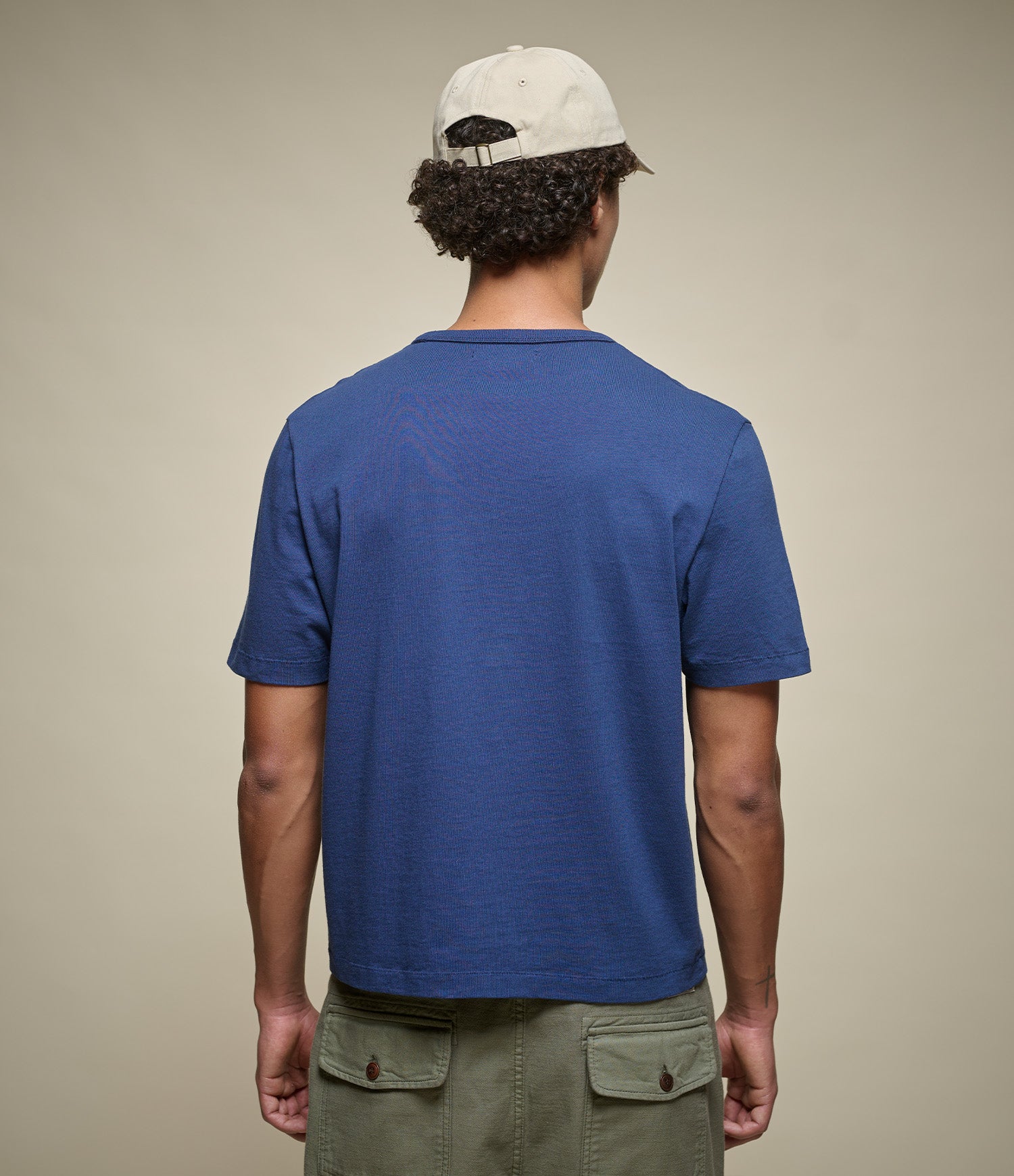 Man wearing a cropped blue t-shirt and beige cap from behind on a plain background