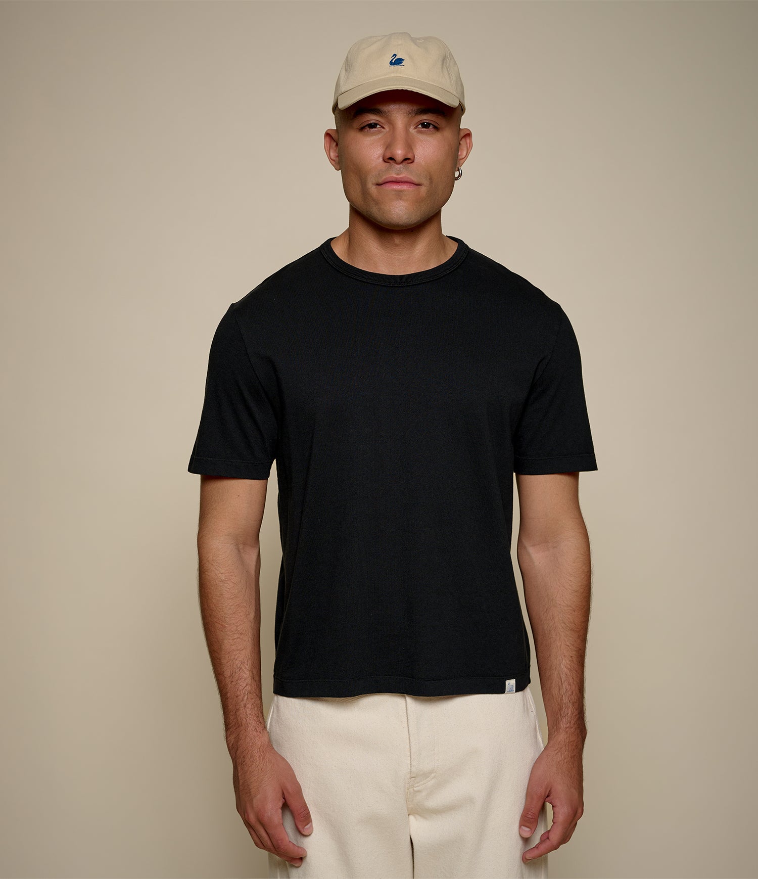 A man wearing a black cropped short-sleeve crewneck t-shirt, cream pants, and a beige baseball cap, standing against a solid beige background.