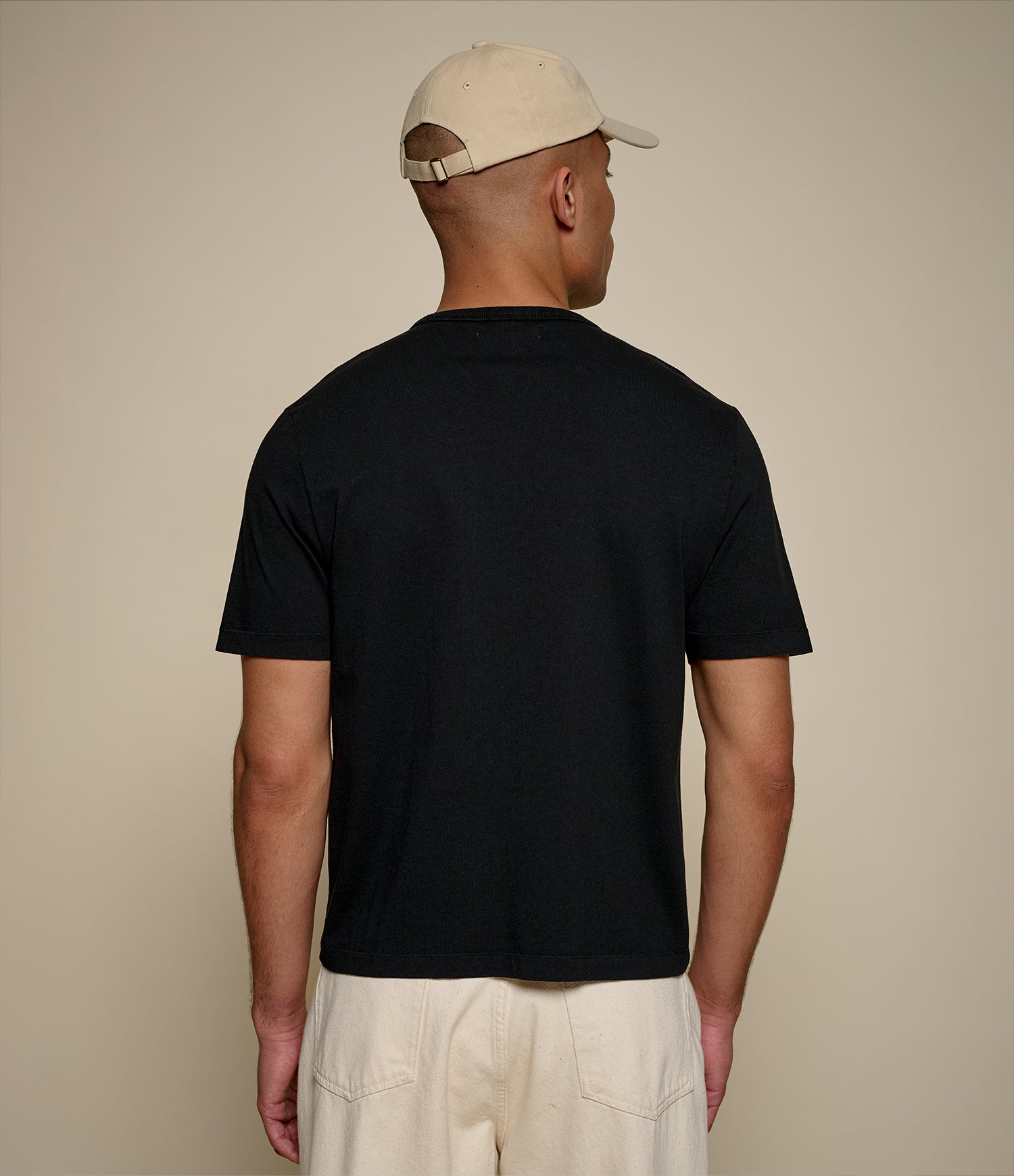 A man viewed from behind wearing a plain black short-sleeve t-shirt, cream-colored shorts, and a cream baseball cap against a solid beige background.