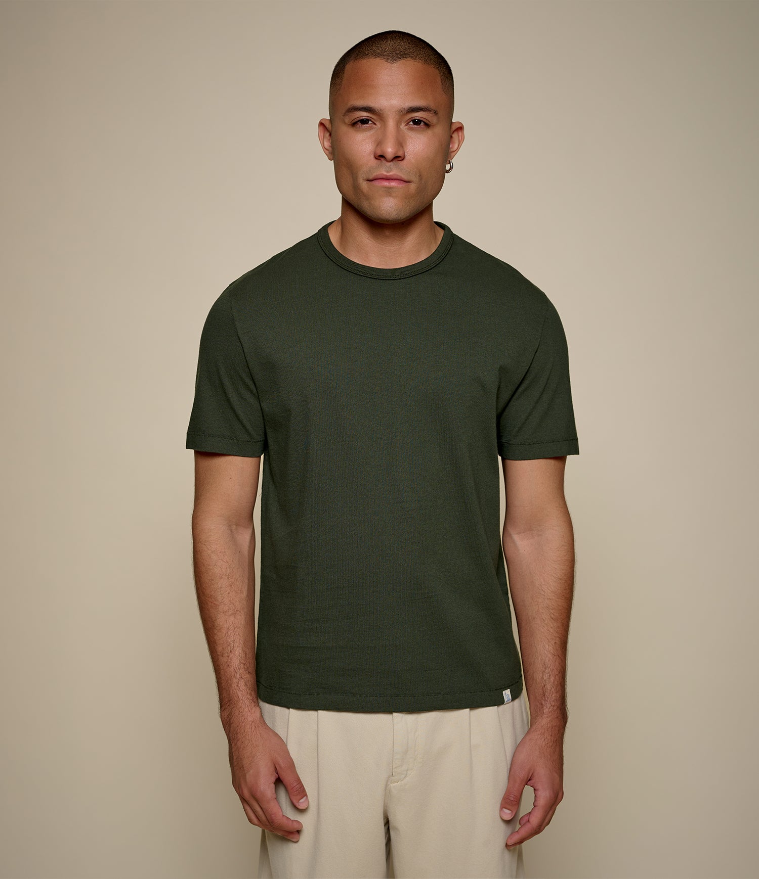 A man wearing a dark olive green crew-neck t-shirt and light beige pants, standing against a solid beige studio background.
