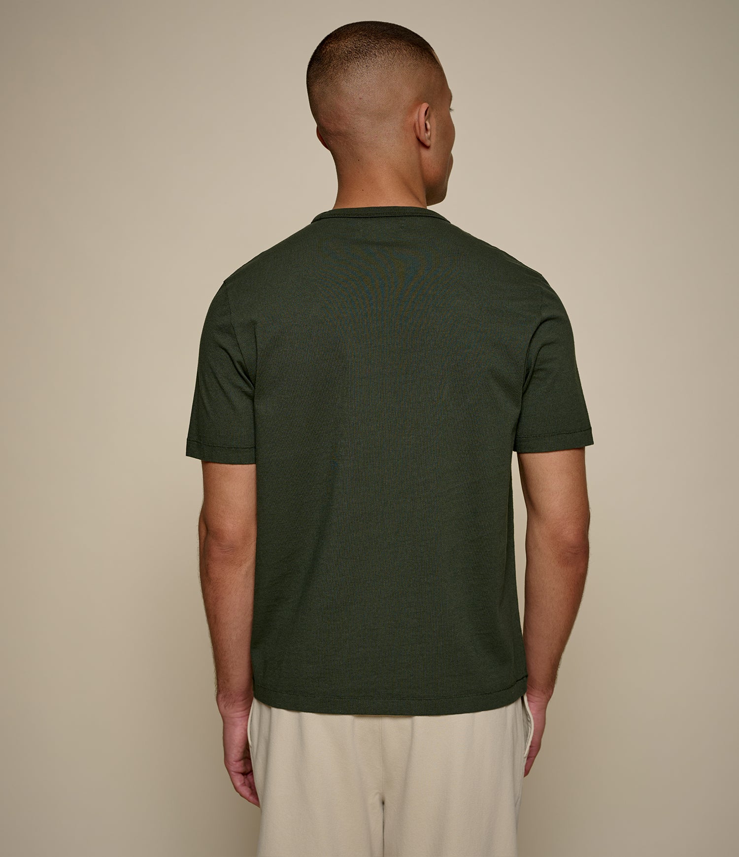 A man viewed from the back wearing a dark olive green short-sleeve t-shirt and light beige trousers, standing against a plain beige studio background.