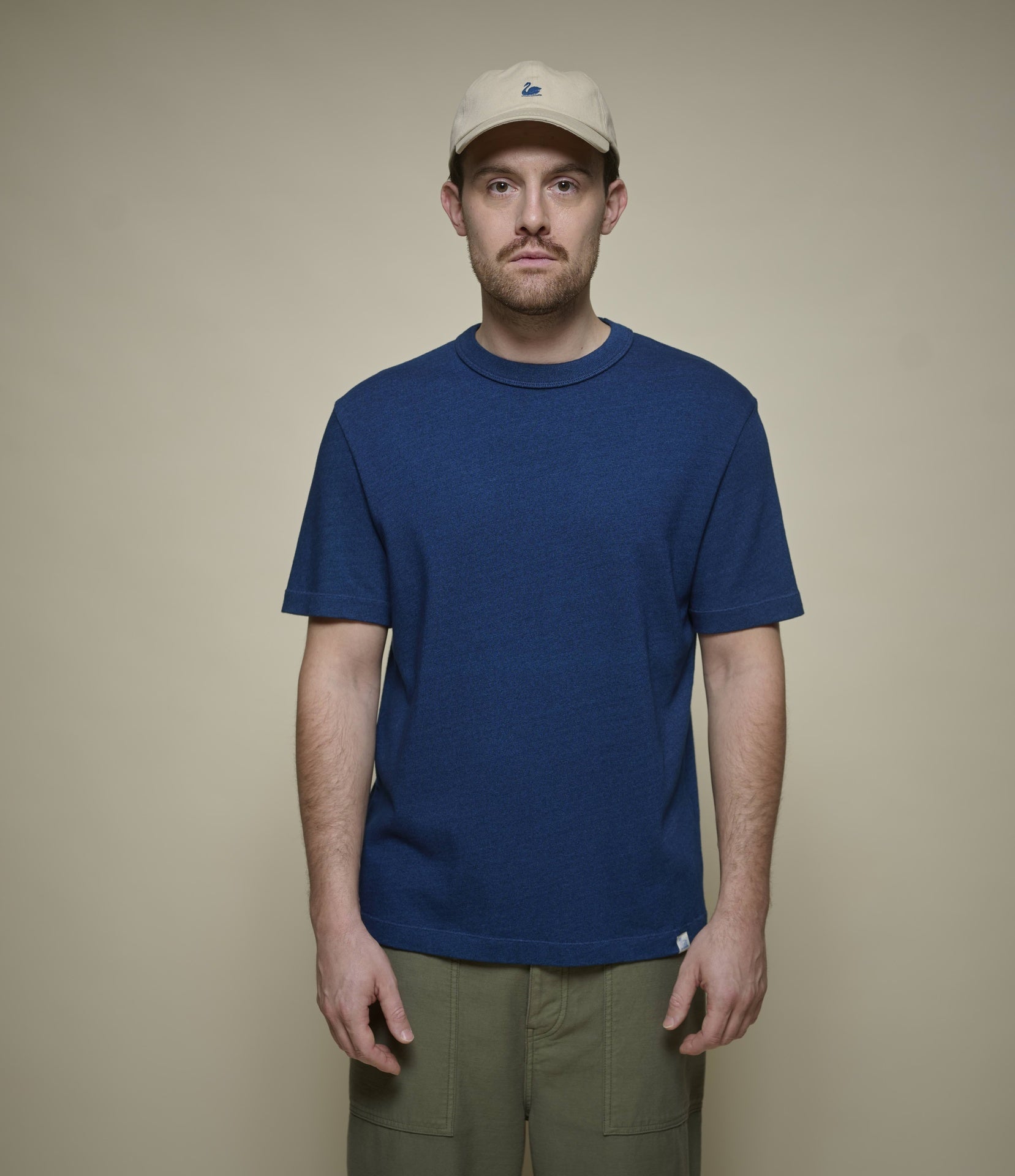 Man wearing an indigo blue authentic jersey t-shirt and beige cap against a beige background
