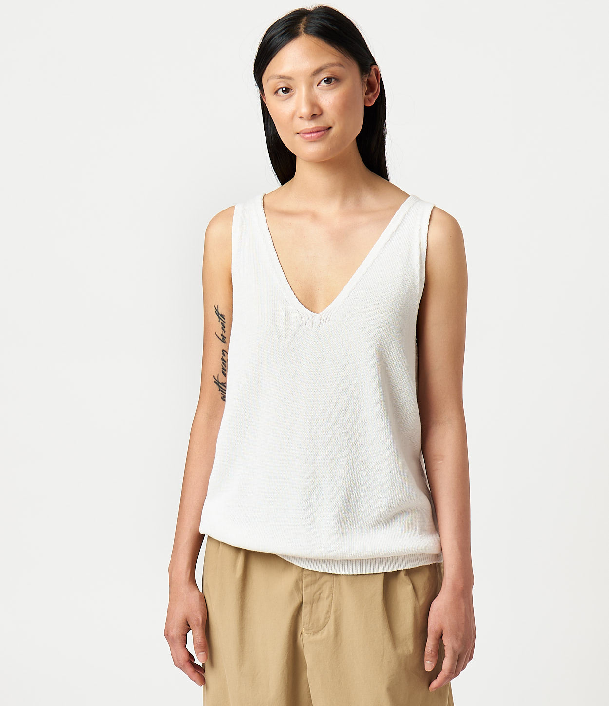 A female model wearing a white knitted top with a relaxed fit, featuring a V-neckline and sleeveless design.